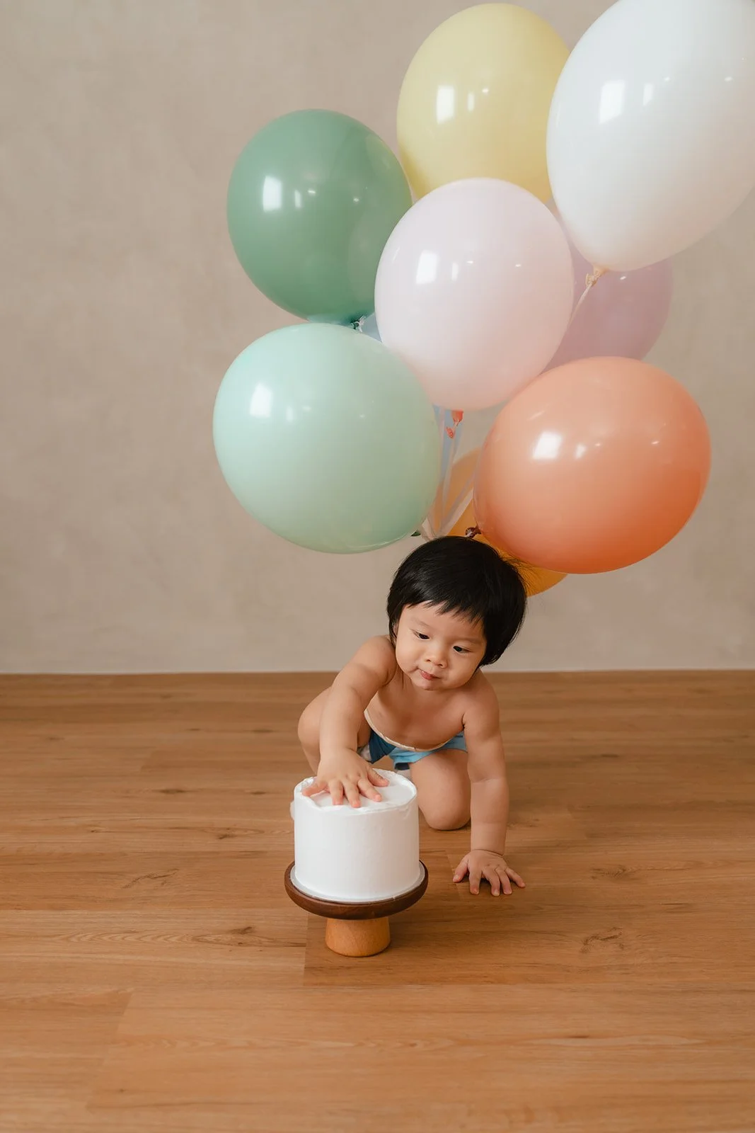 Child in underwear reaching for a small white cake with a wooden cake stand, surrounded by pastel-colored balloons, on a wooden floor.