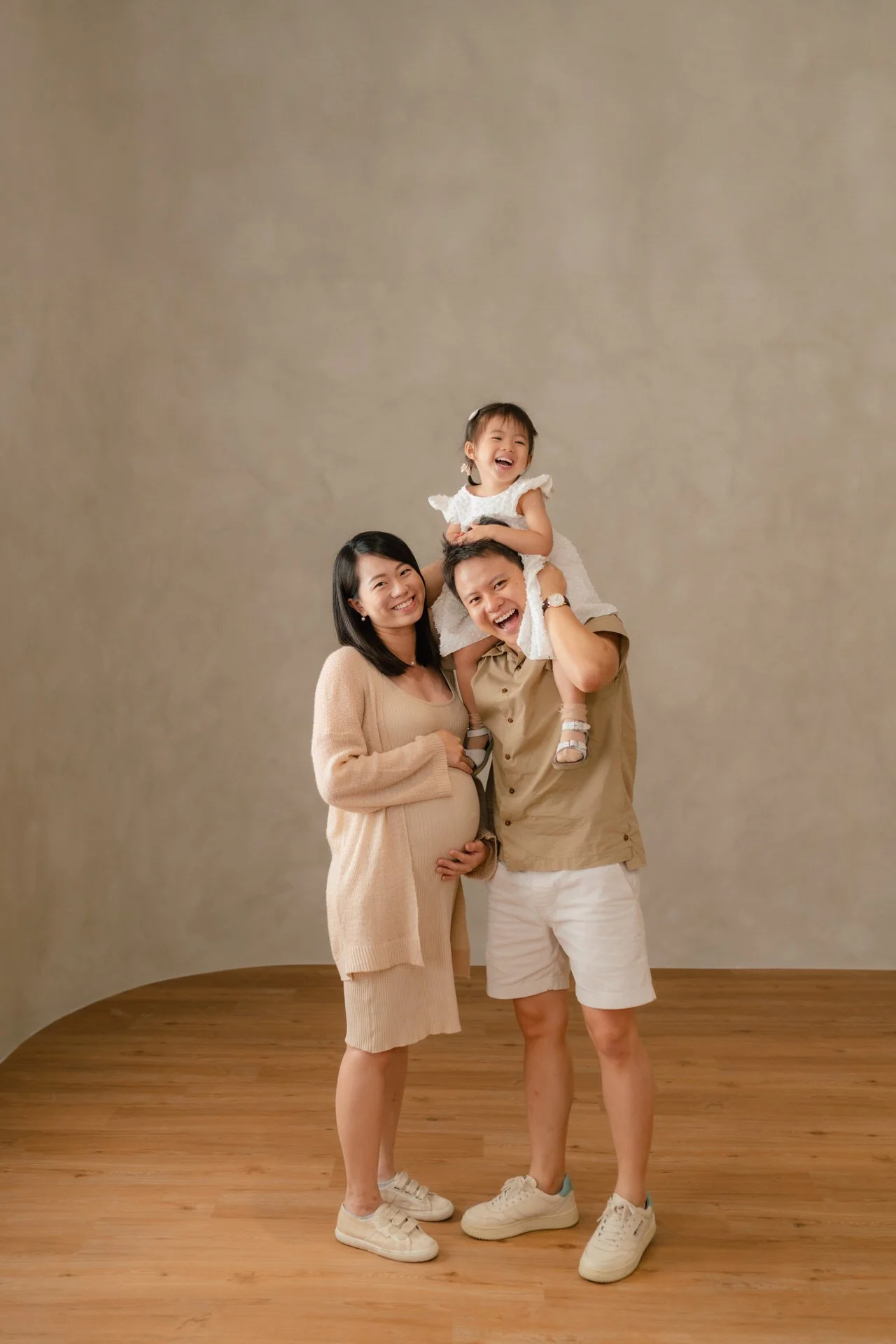 Happy family consisting of a pregnant woman, a man, and a young girl, standing on a wooden floor against a plain background. The girl is sitting on the man's shoulders, and the family is smiling and looking at the camera.