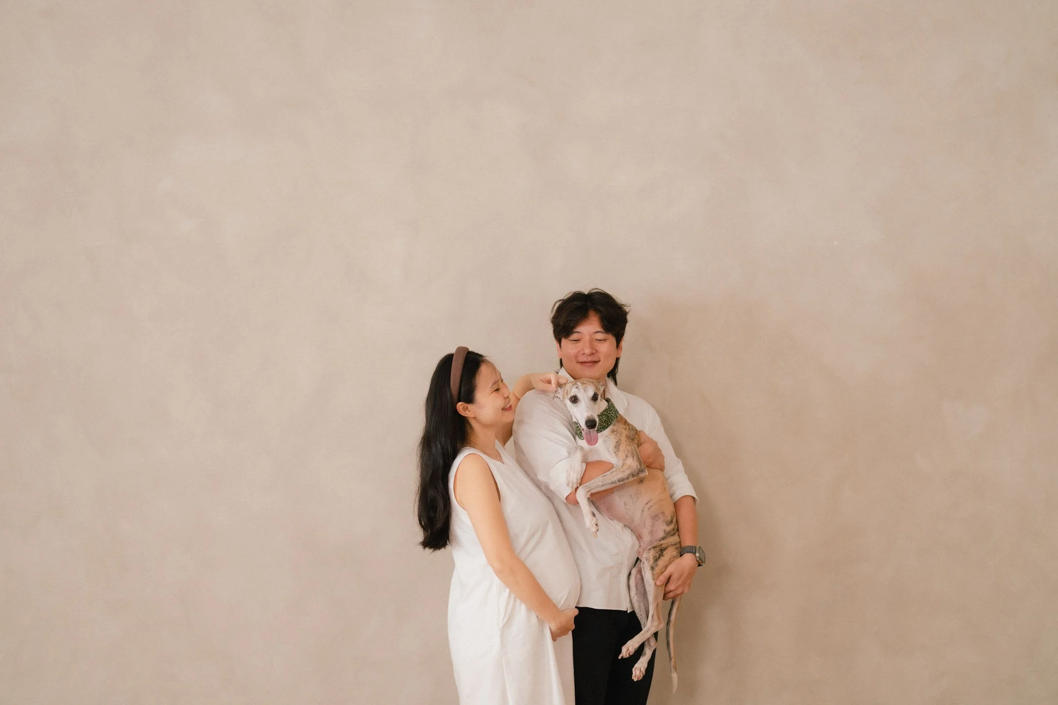 A woman and a man standing close together, with the woman smiling and the man holding a dog and smiling, against a plain beige wall.