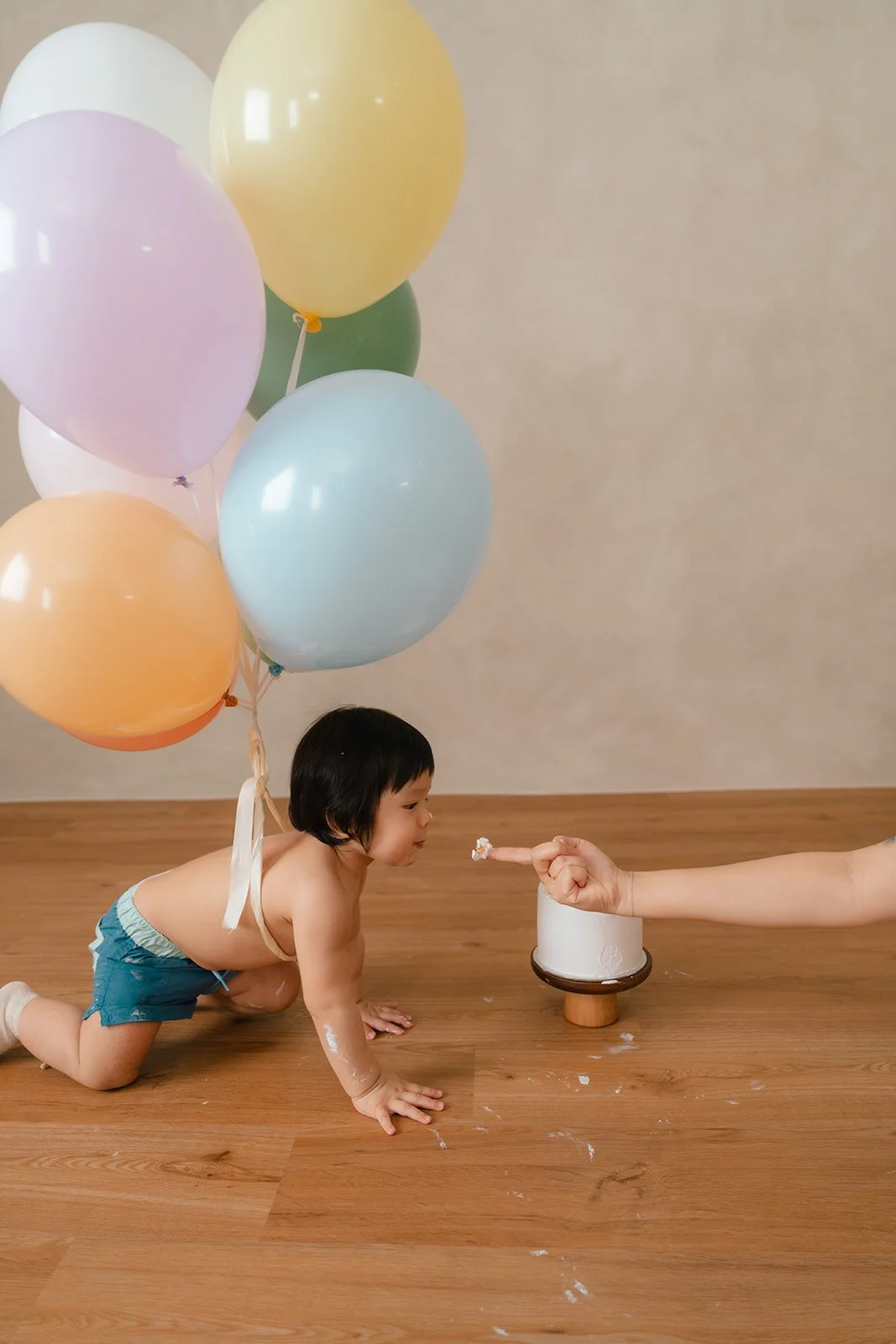 A young child in blue shorts and no shirt is crawling on a wooden floor towards a small cake, with a person’s arm pointing a finger at the cake. There are pastel-colored balloons floating in the air behind the child, suggesting a celebration.
