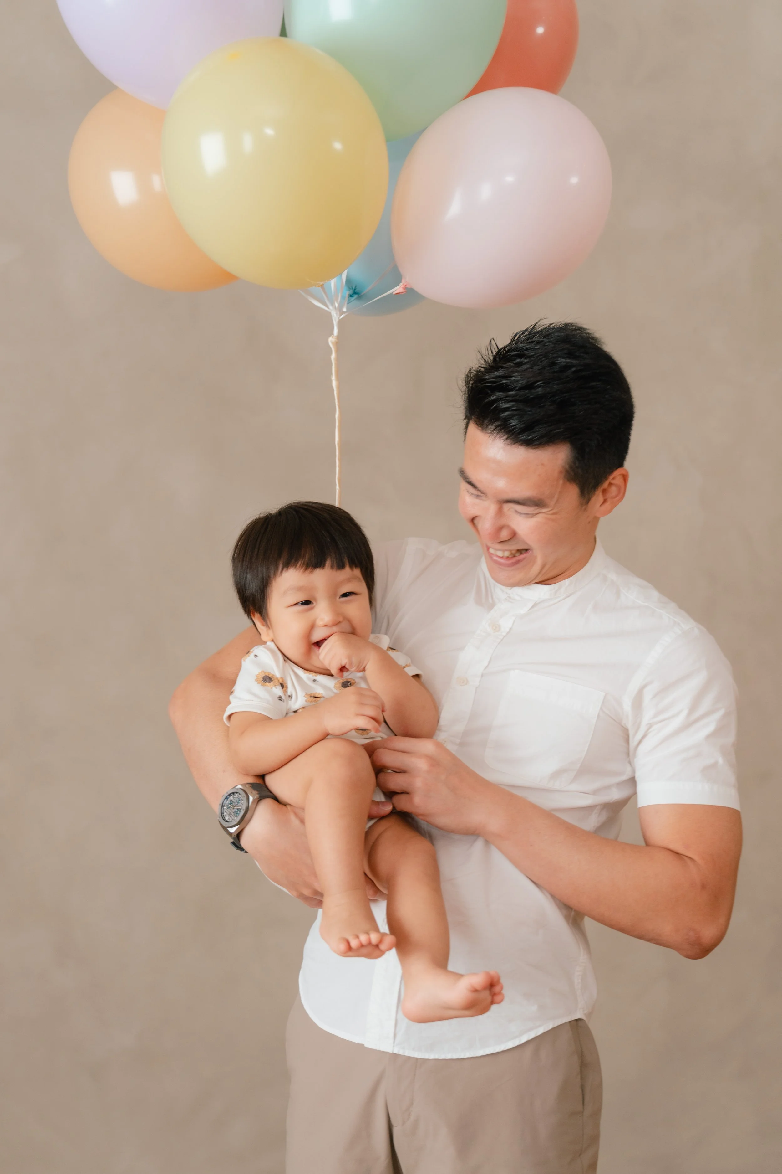A man holding a young child while smiling, with pastel-colored balloons floating above them, celebrating a birthday.