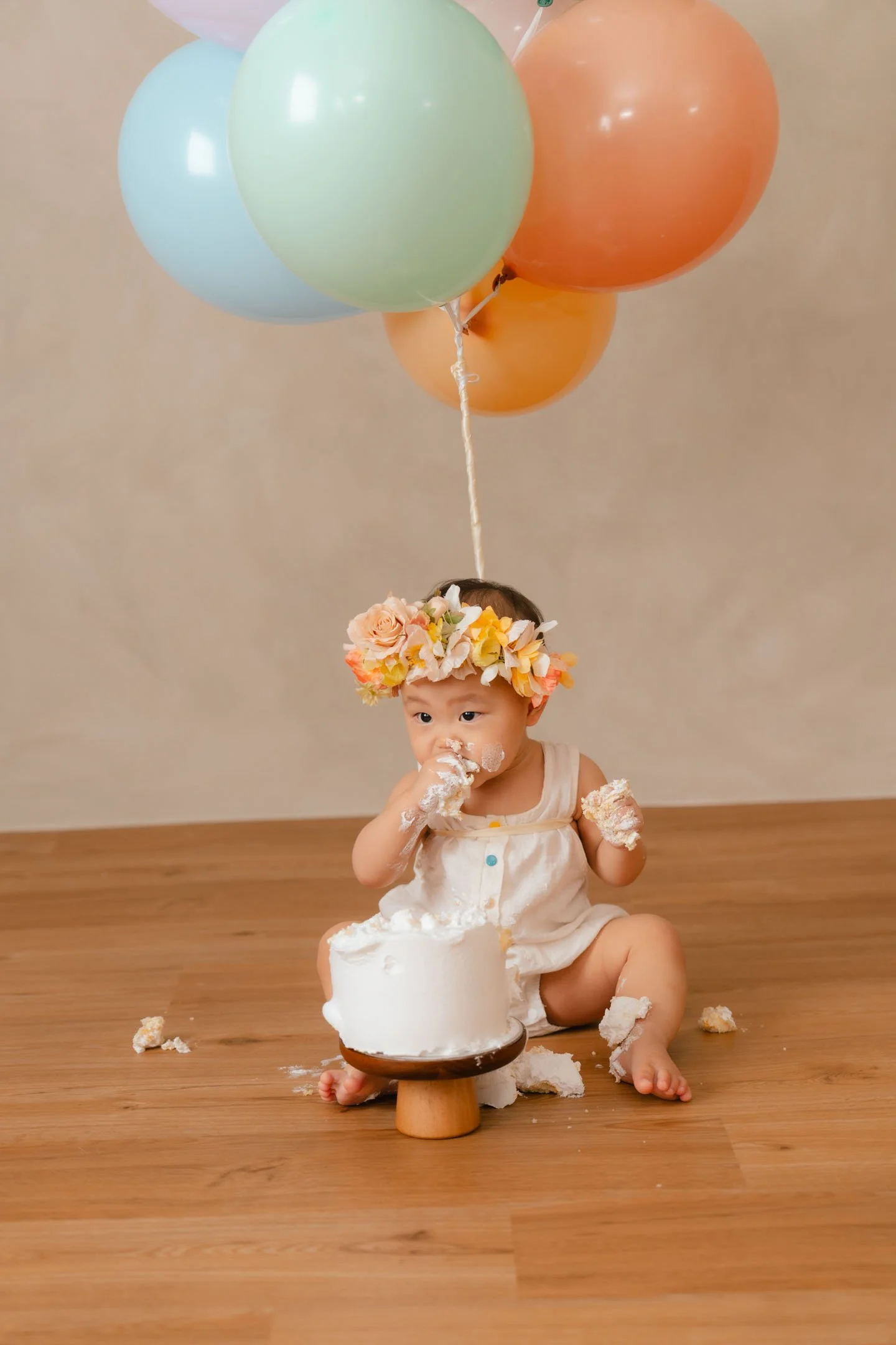 A young child with a flower crown celebrating a birthday, sitting on a wooden floor in front of a cake with her hand in frosting, surrounded by balloons.