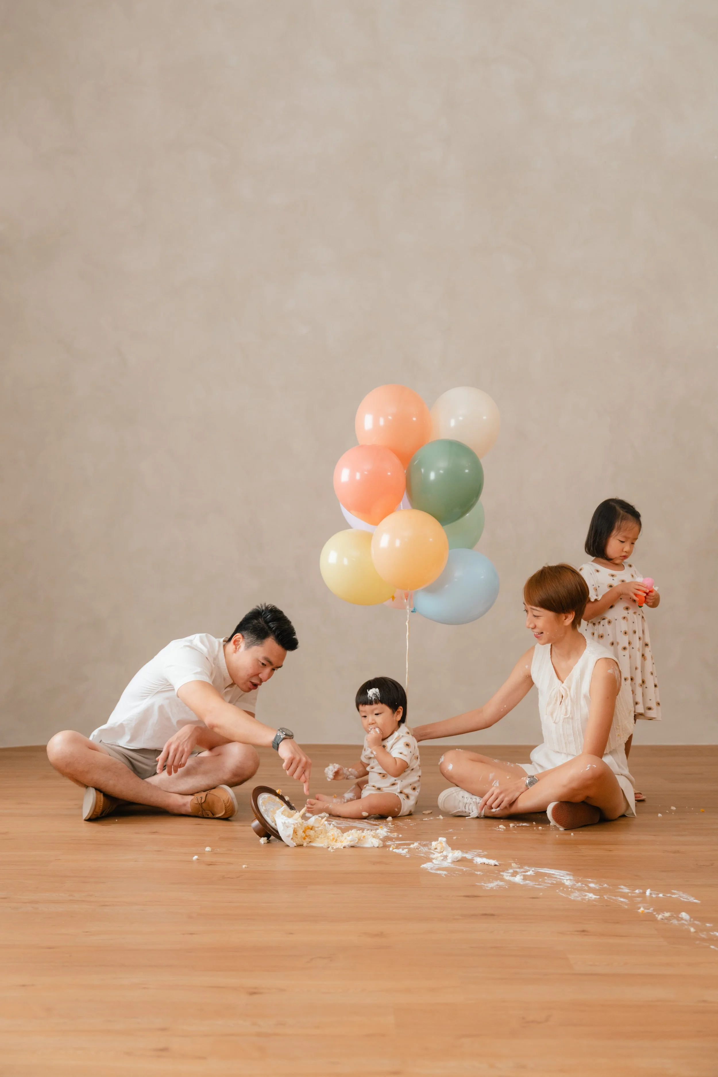 A family of four celebrating a child's birthday with balloons and cake, with two children and two adults sitting on a wooden floor, one child standing, and colorful balloons in the background.