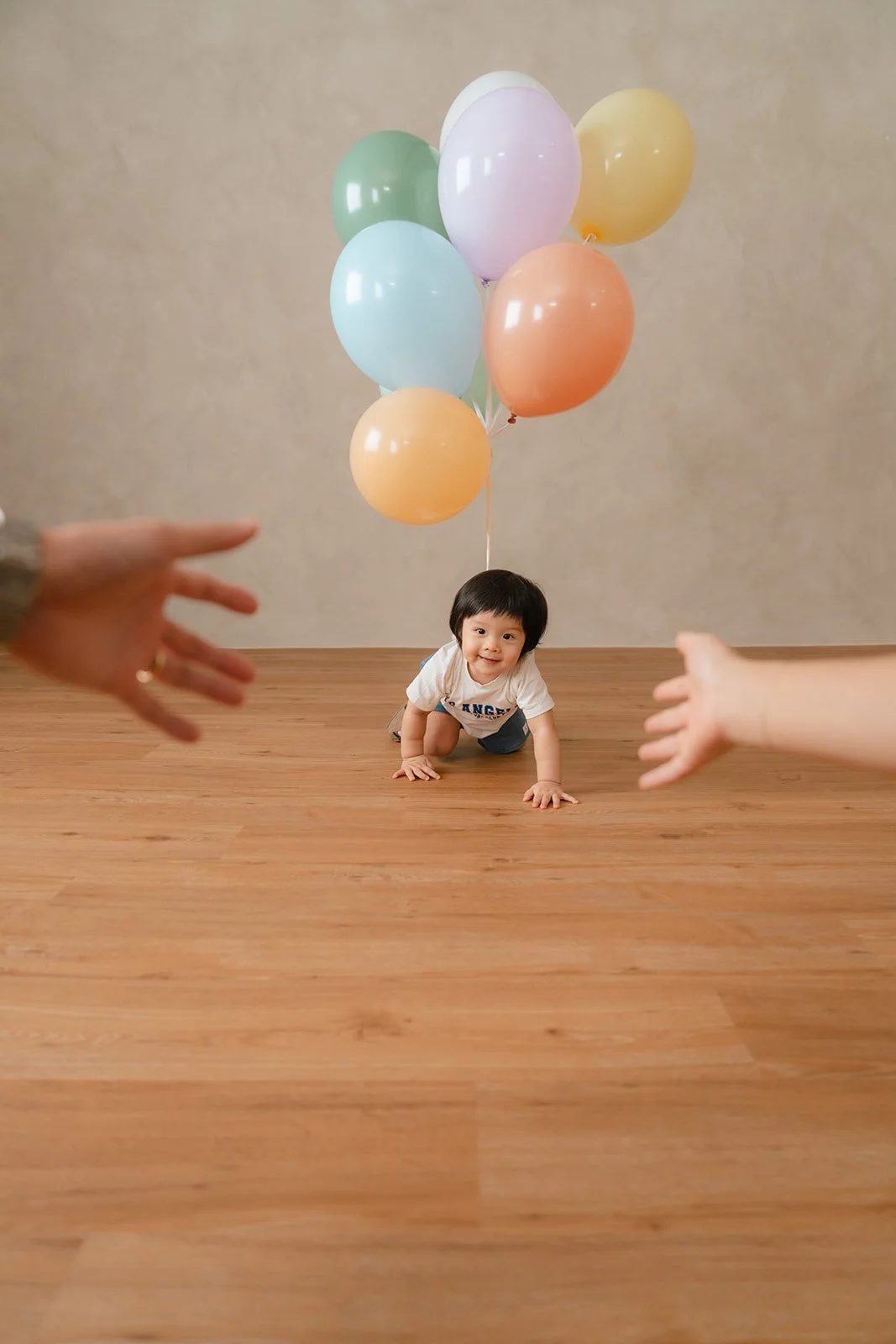 A young child crawling on a wooden floor, holding a bunch of pastel-colored balloons behind them, with adult hands reaching toward the child from the sides.