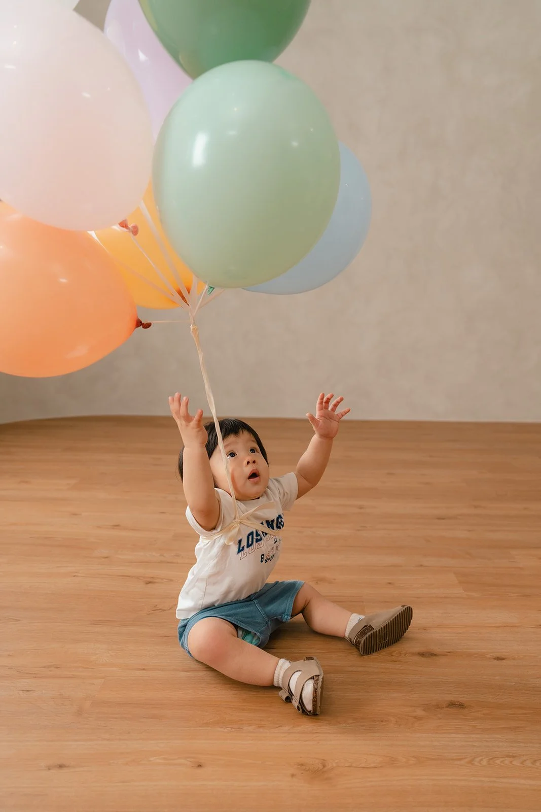 A young child sitting on a wooden floor reaching up towards pastel-colored balloons floating above him.