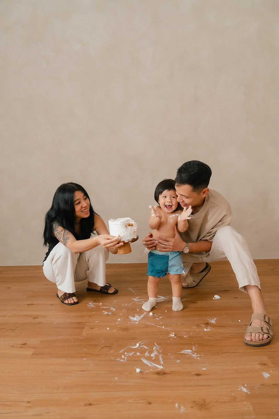 A family celebrating a child's birthday with cake, a woman holding the cake, a smiling toddler covered in cake, and a man crouching next to the child, on a wooden floor with cake mess.