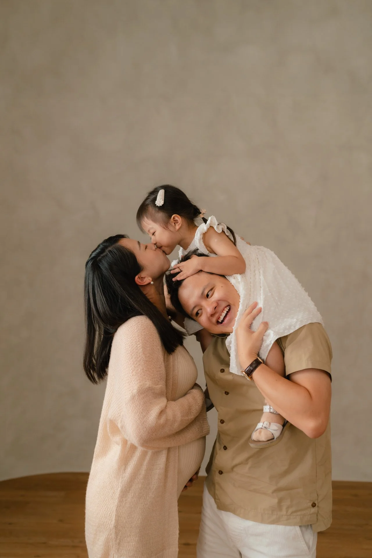 A family of three celebrating a pregnancy, with the mother, father, and young daughter sharing a moment of love, as the daughter kisses her mother.