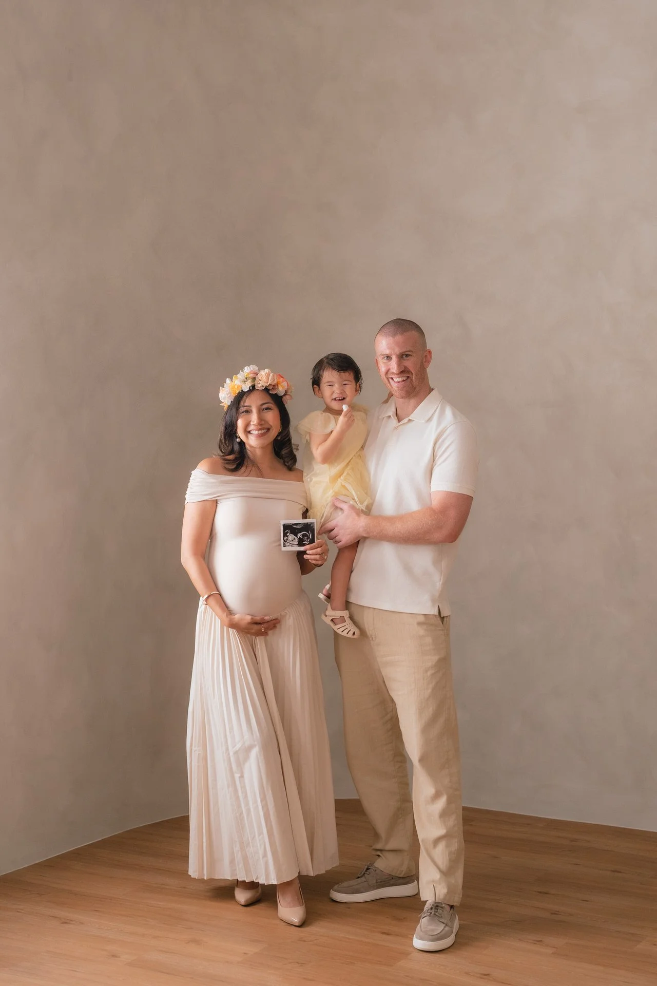 A pregnant woman wearing a flower crown and pleated dress standing next to a man holding a little girl. The woman is holding an ultrasound photo. They are all smiling and dressed in light-colored clothing, standing on a wooden floor with a plain back
