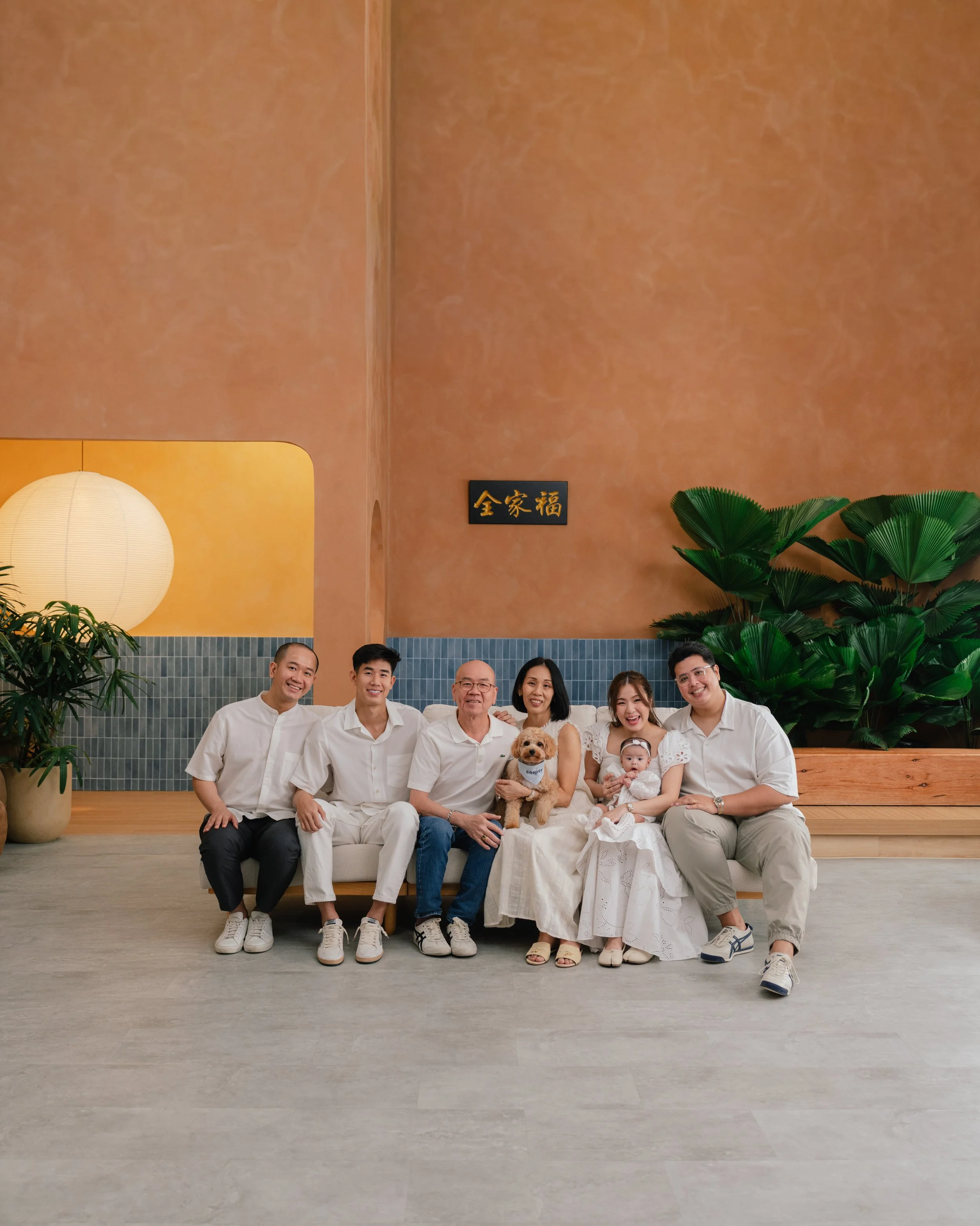 A group of eight people, including children and adults, sitting on a white round sofa in a modern, minimalistic room with a large potted tree, wooden accents, and beige walls, all smiling and laughing.