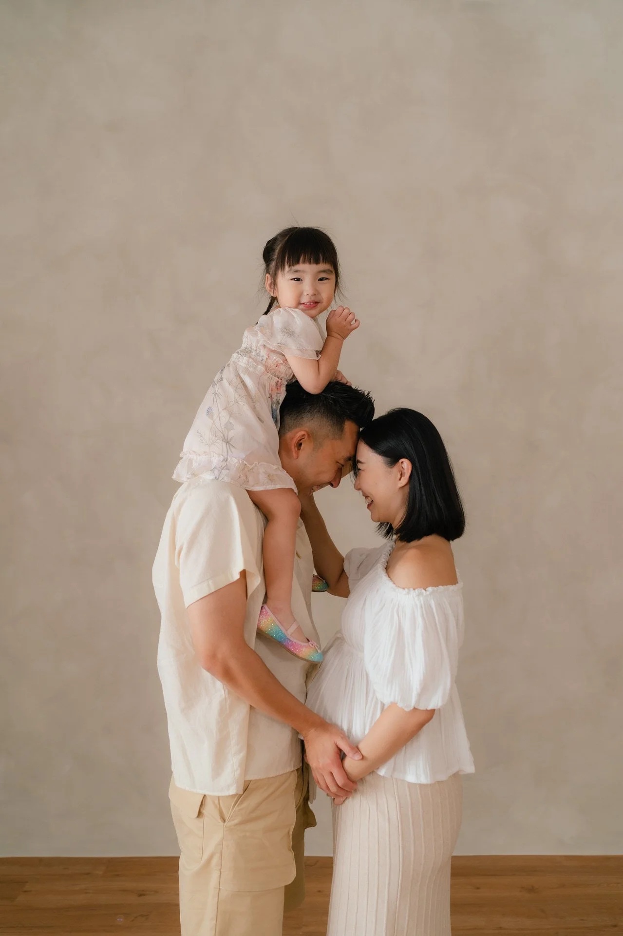 A family of three, including a man, a woman, and a young girl, happy and smiling together. The man and woman are standing close, with the young girl sitting on the man's shoulders and leaning forward to touch foreheads with the woman.