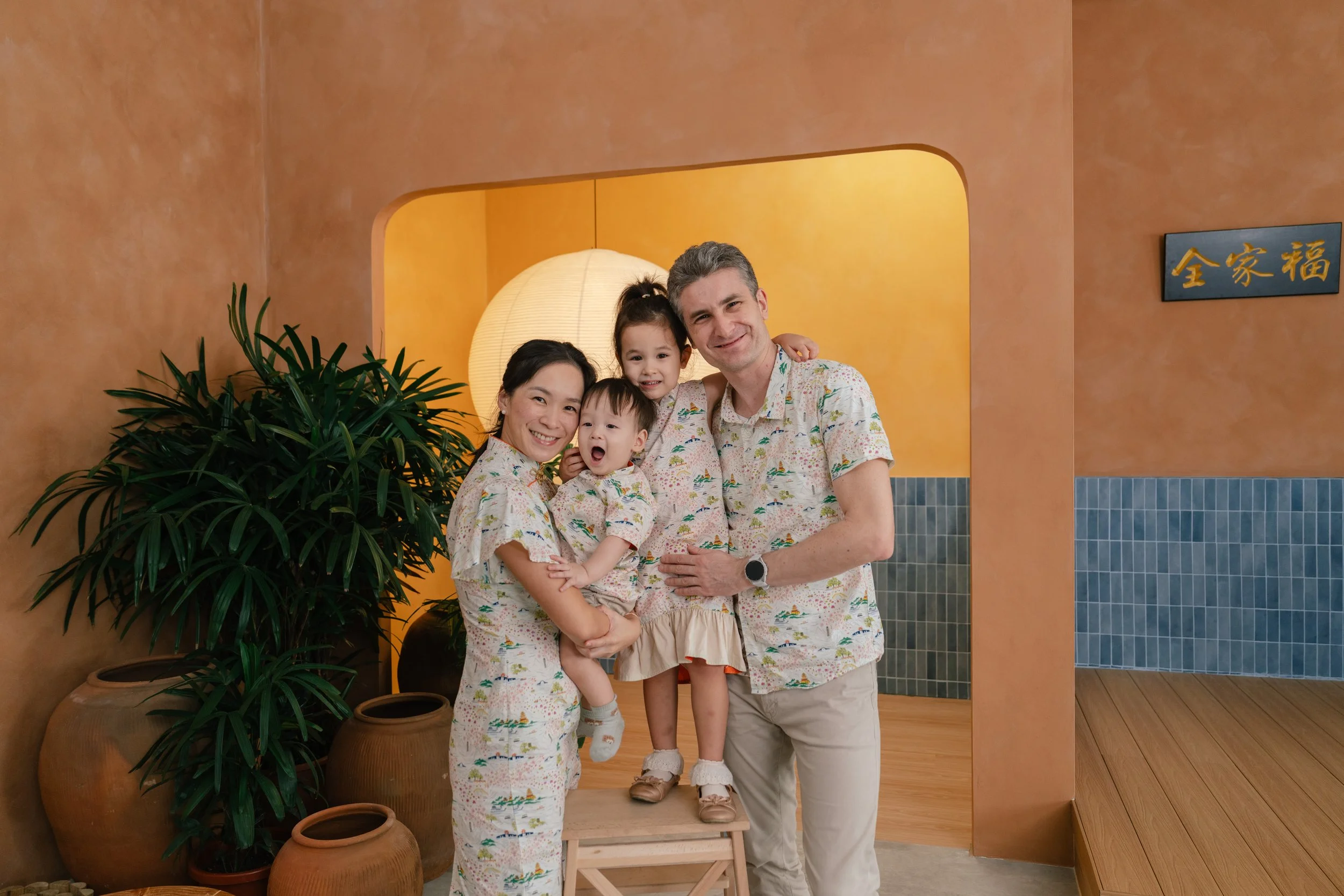 A group of eight smiling people, including children and adults, sitting closely together on a white sofa in a modern, minimalist room with wooden flooring, a large potted plant, and round wooden stools.