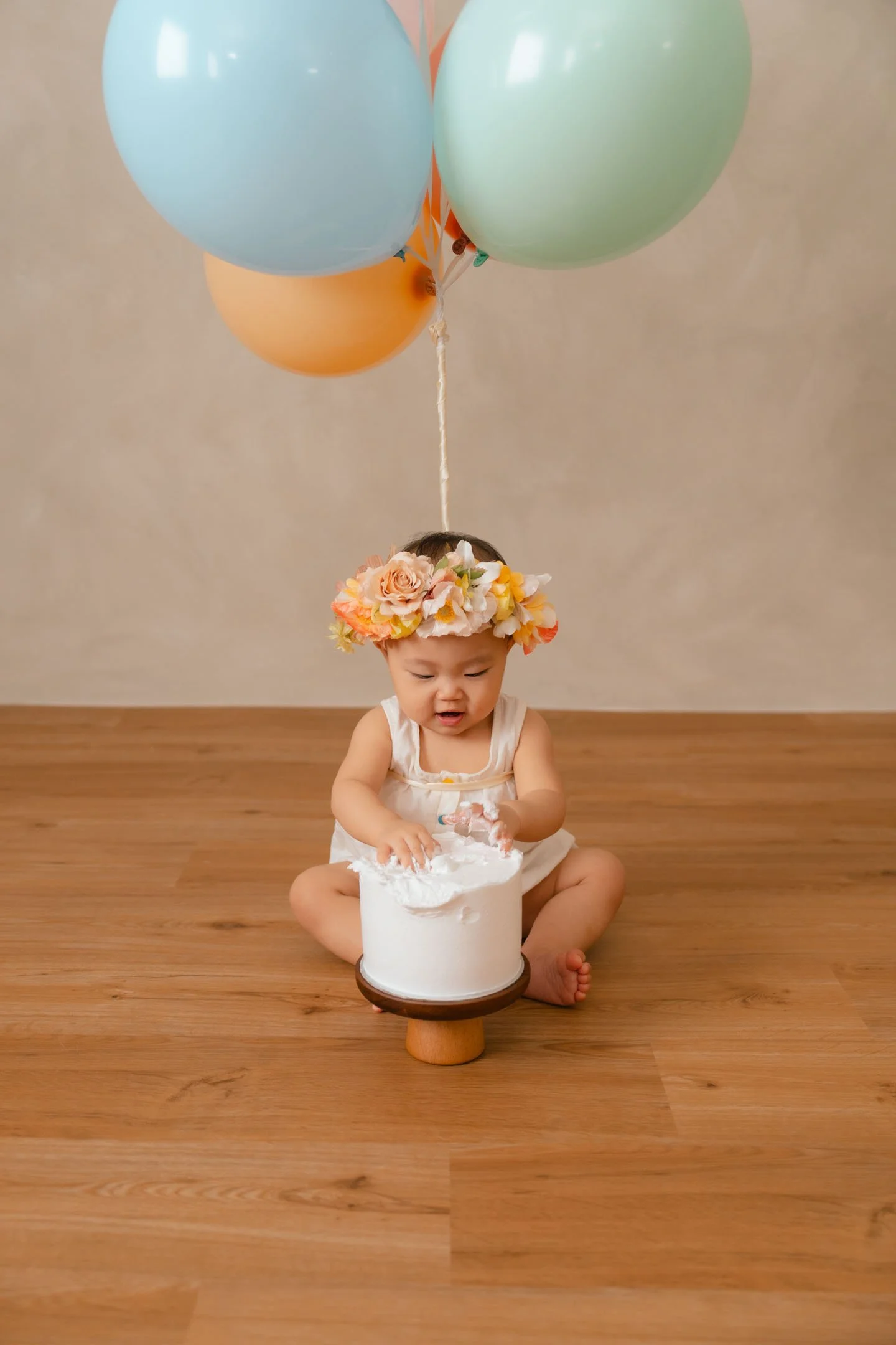 A young child wearing a floral headband and a white dress, sitting on a wooden floor, playing with a small white cake, with three pastel-colored balloons floating above her.