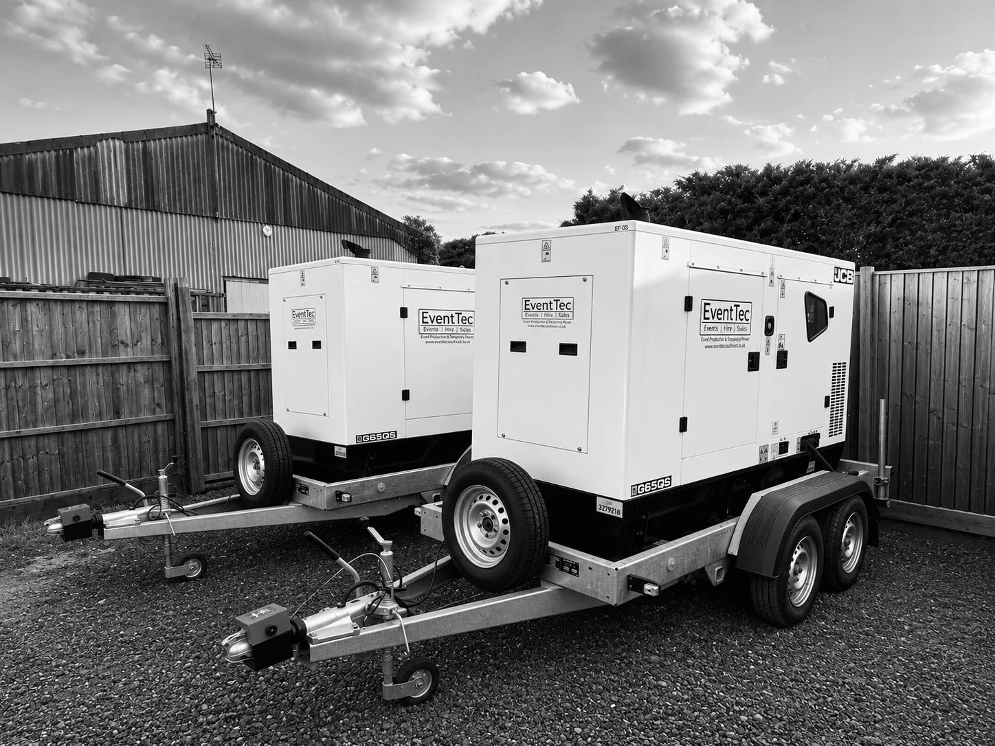Two mobile generators on trailers parked on gravel in an outdoor lot with wooden fences and a large warehouse building in the background. The generators are branded with EventTec and JCB logos, and each is mounted on a dual-axle trailer with wheels and support legs.