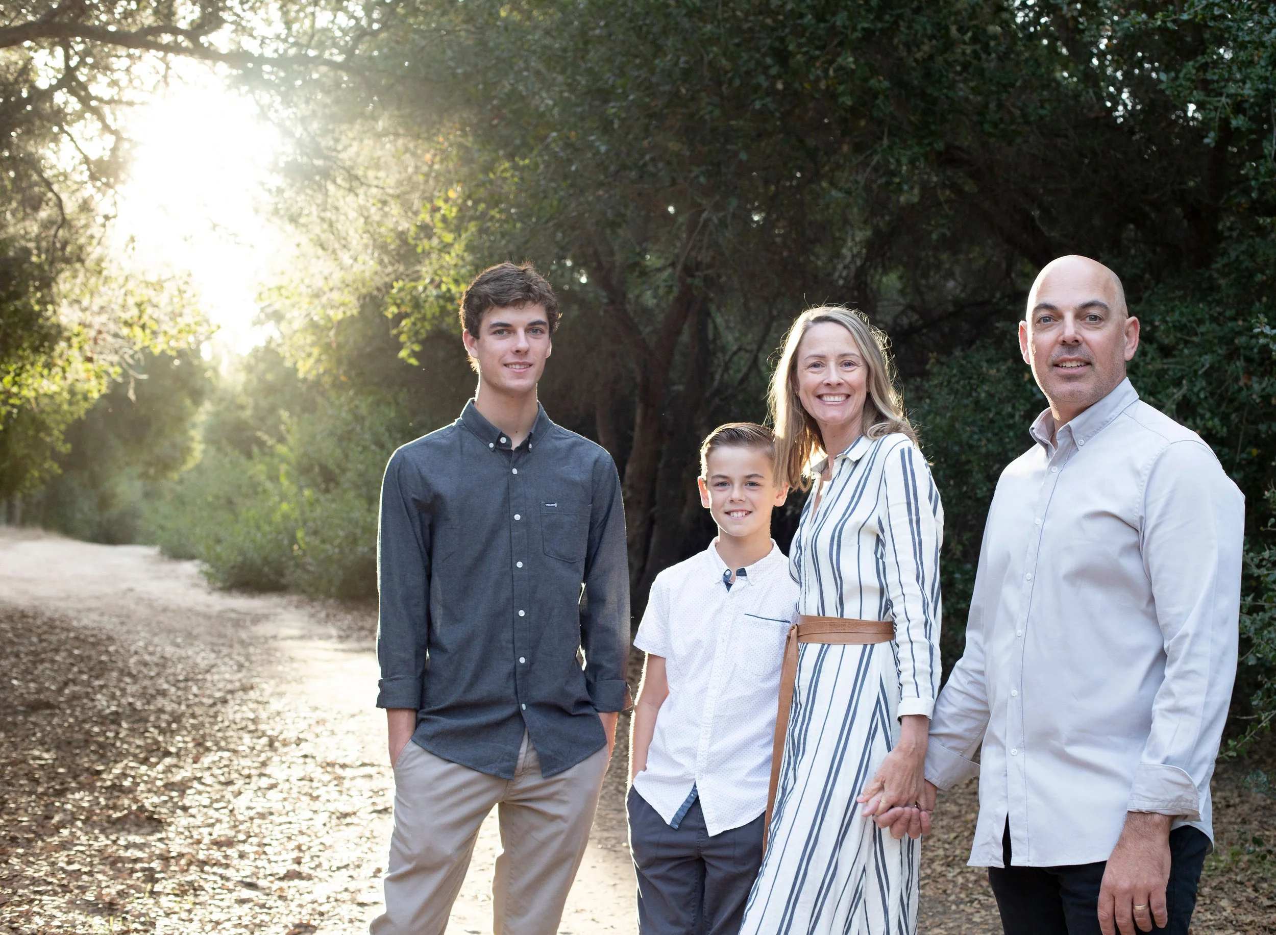Family of five standing on a dirt path in a wooded area, holding hands and smiling, with sunlight filtering through the trees.