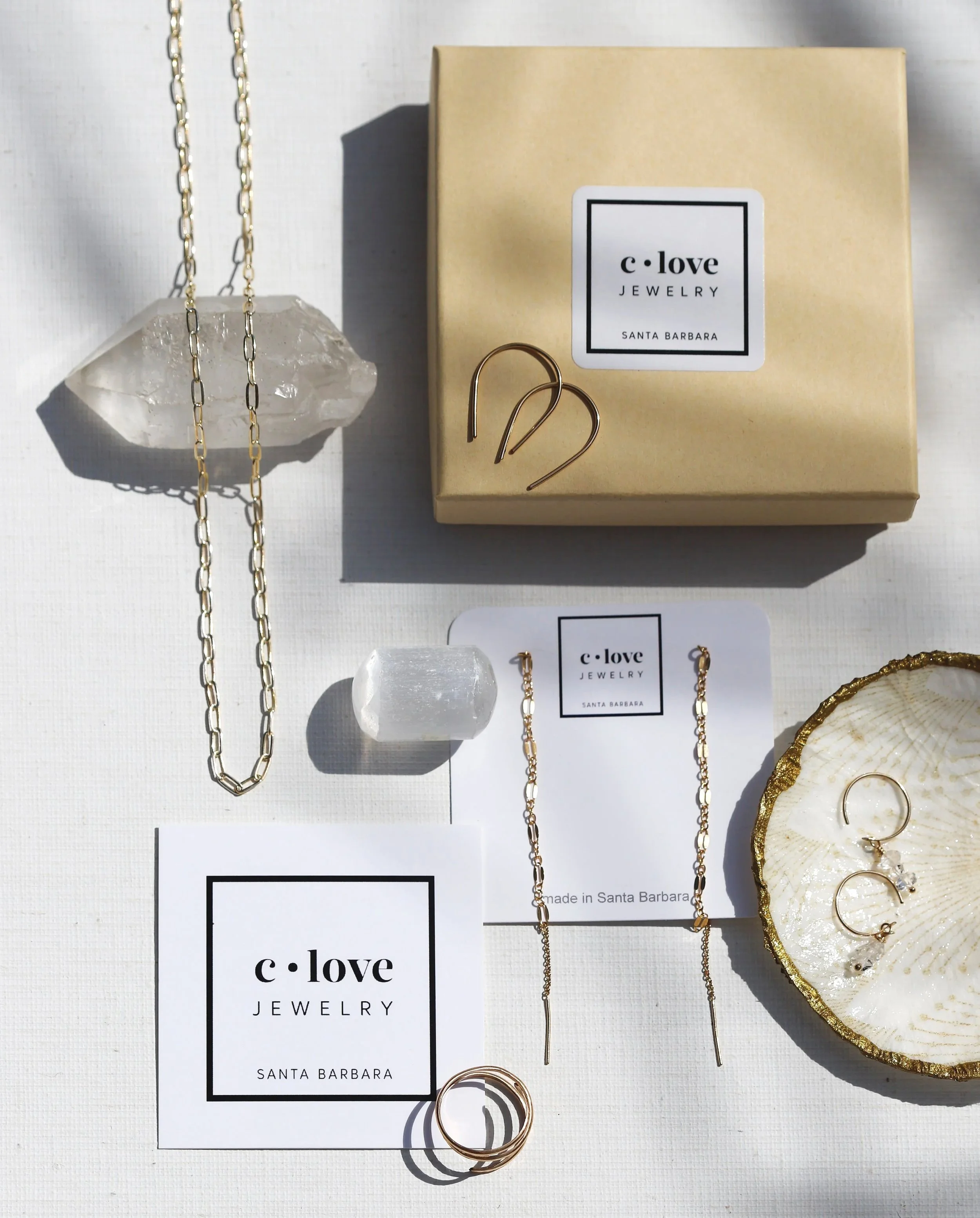 Display of jewelry including a silver chain, gold earrings, a gold ring, and a gold bracelet on a white surface with a jewelry box labeled 'c-love Jewelry Santa Barbara'.