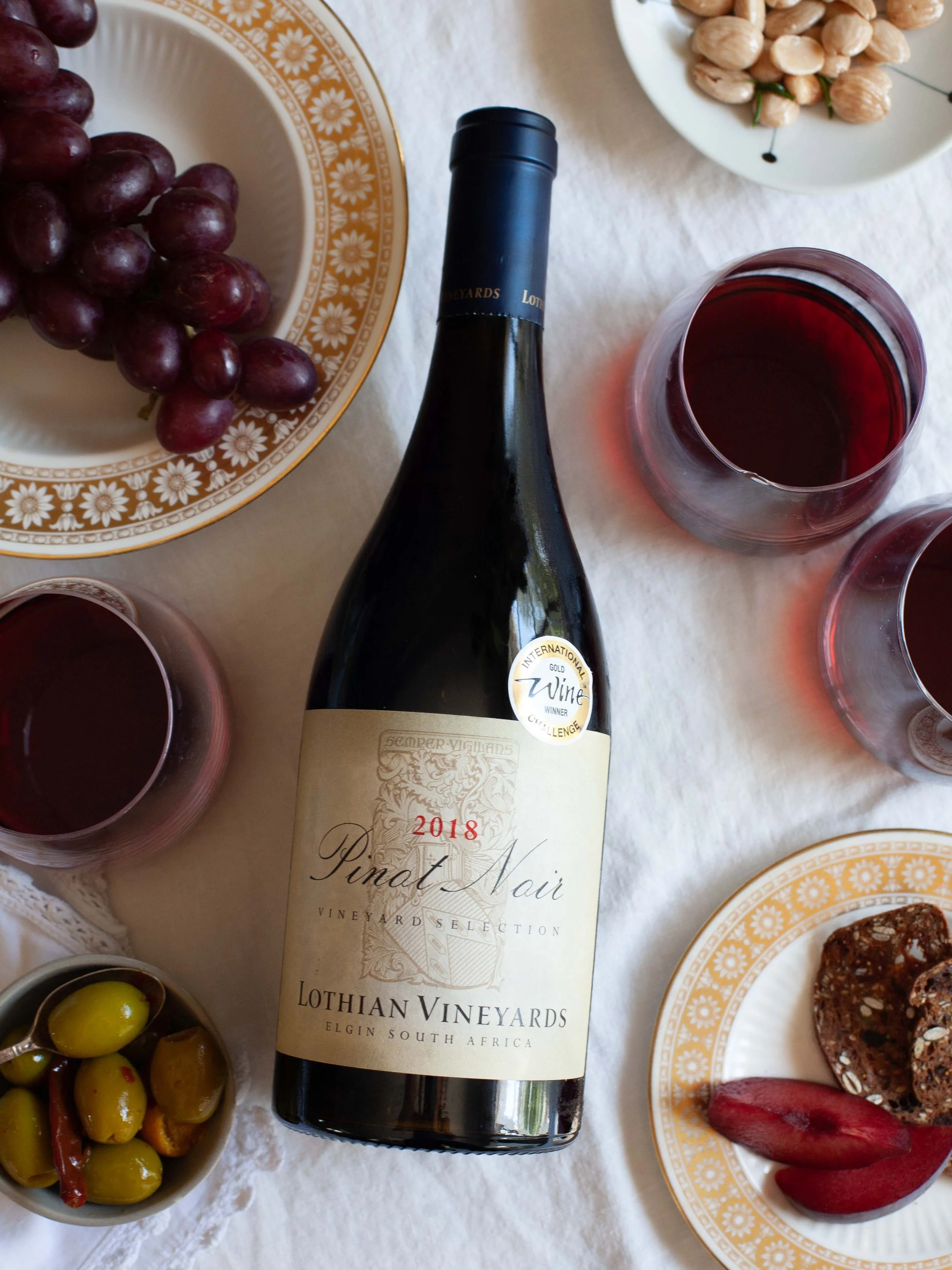 A bottle of Lothian Vineyards Pinot Noir wine on a table alongside plates of grapes, olives, and assorted foods.