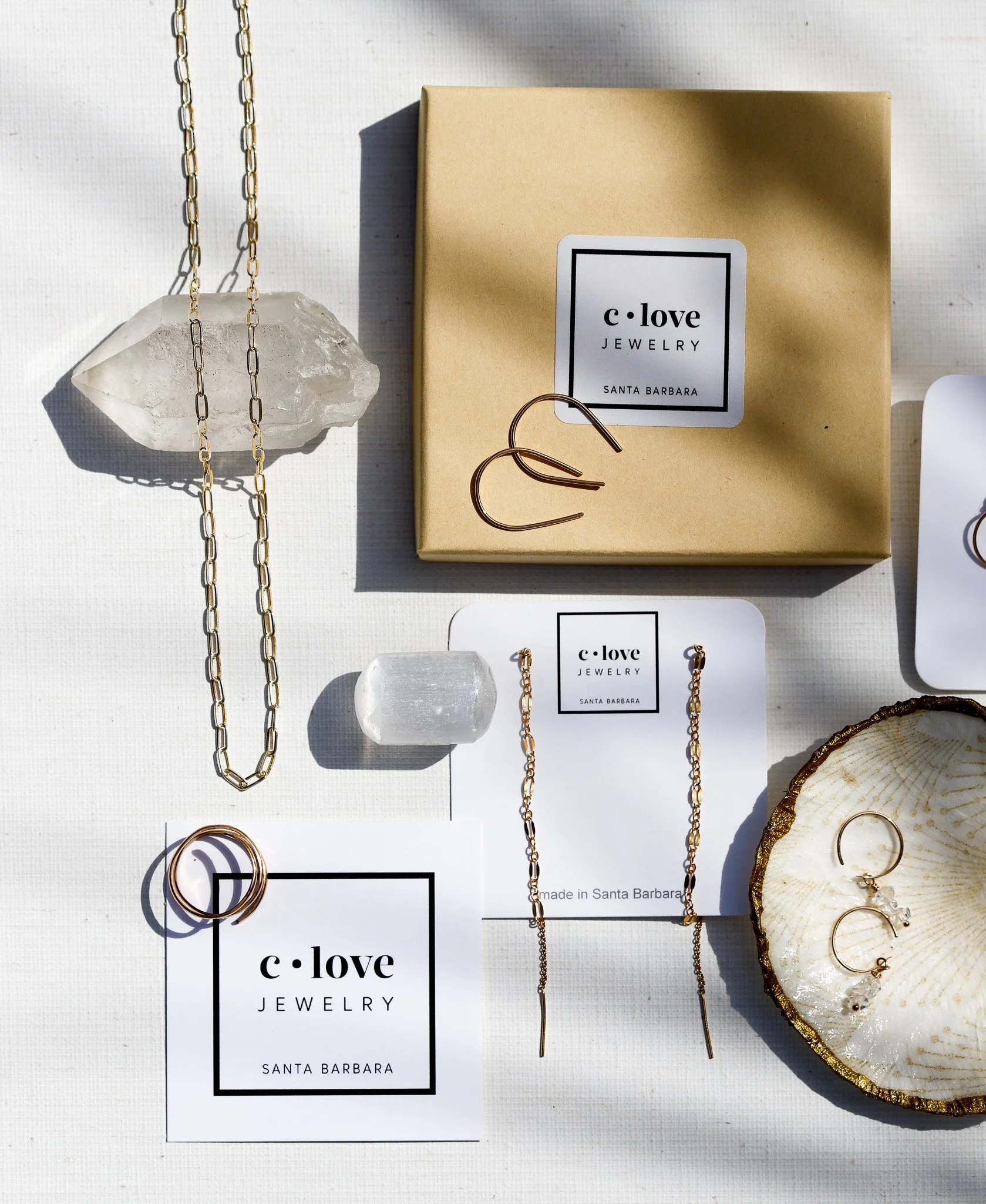 Jewelry display featuring gold chains, rings, and earrings on a white surface, with a branded jewelry tag from 'c love Jewelry Santa Barbara' and a decorative shell with gold trim.