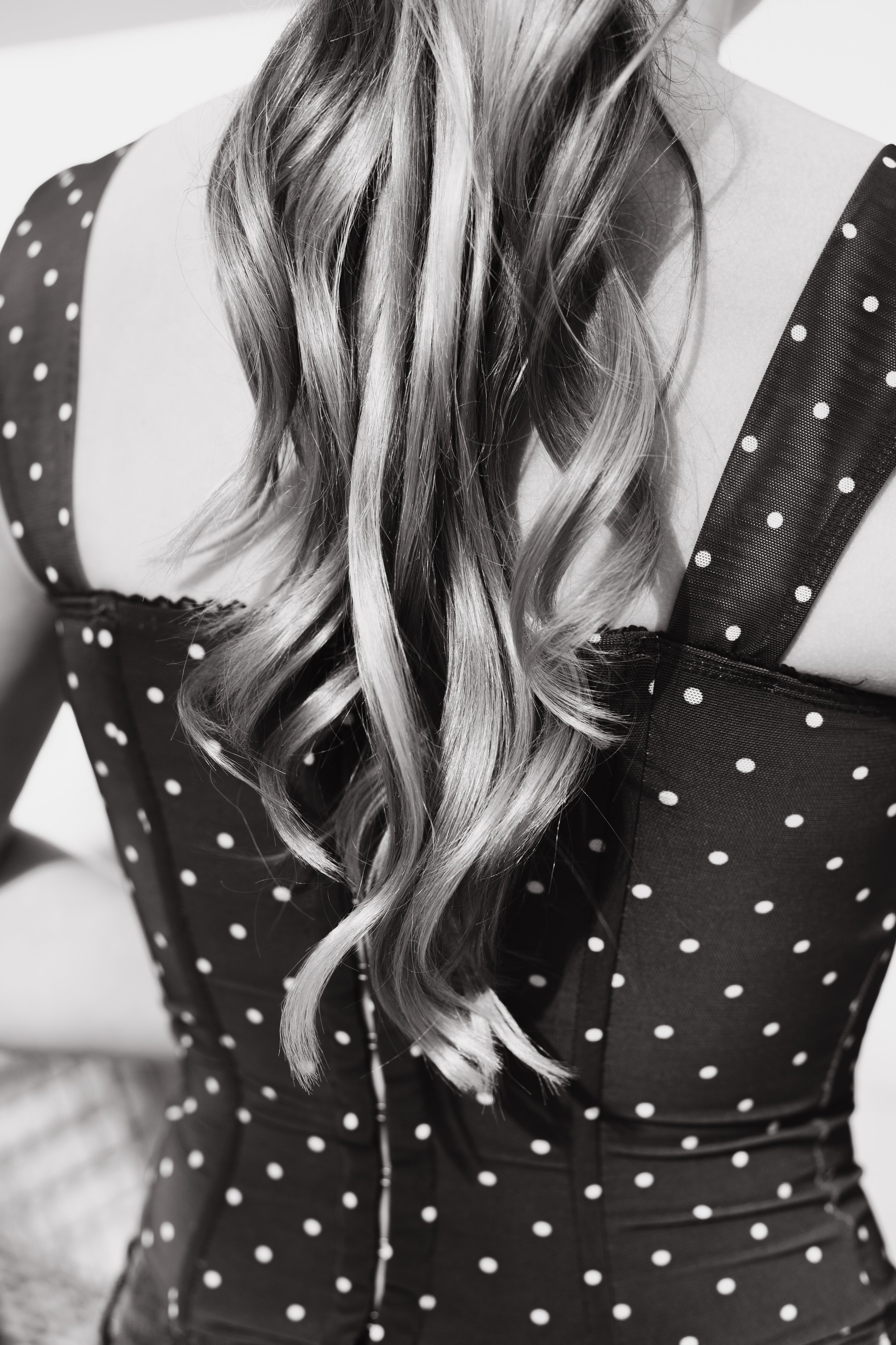 Close-up of a woman's shoulder and long wavy hair, wearing a polka-dot dress with a zipper at the front, black and white image.
