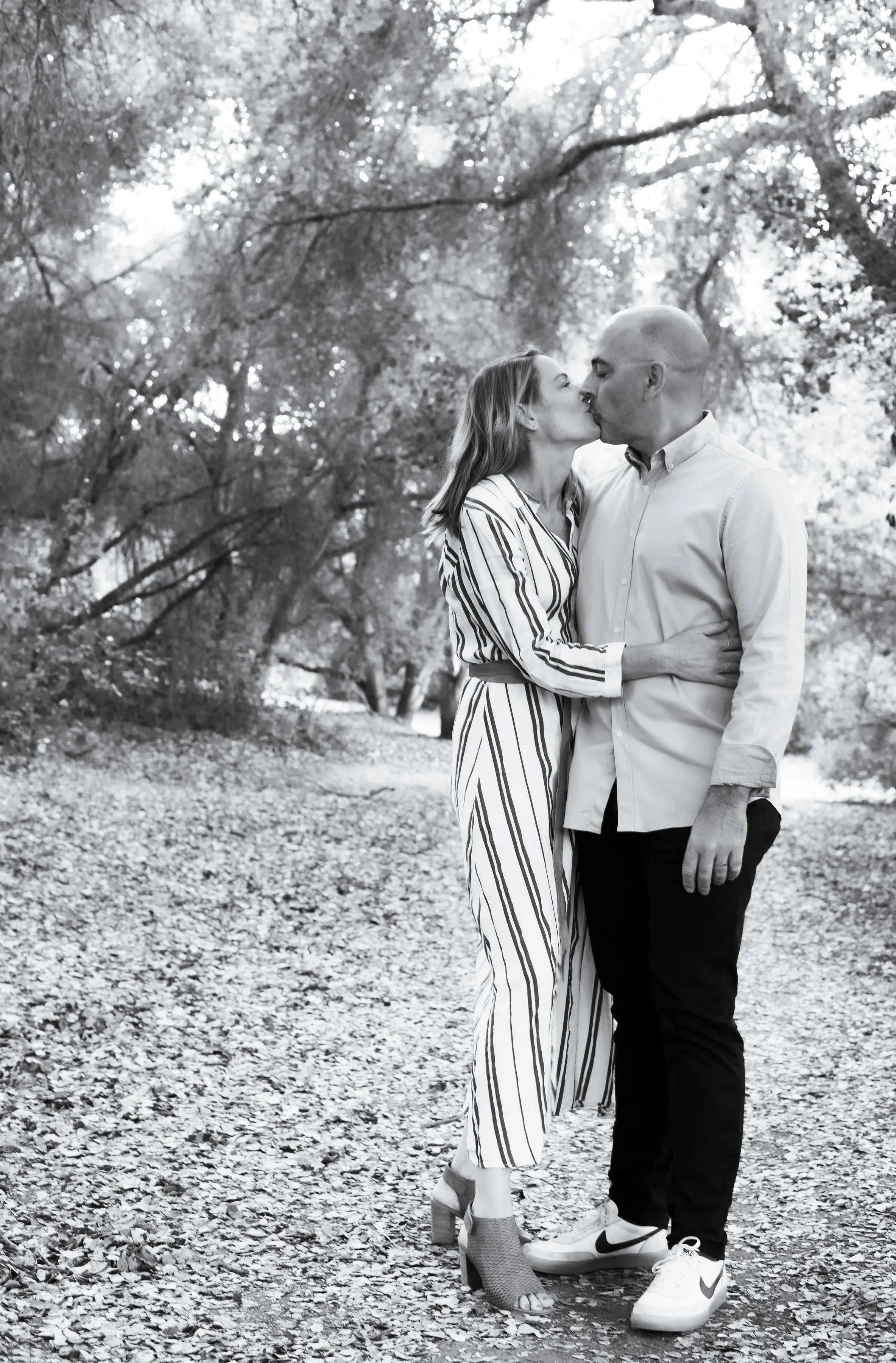 A couple kissing outdoors on a leaf-covered path surrounded by trees in black and white.