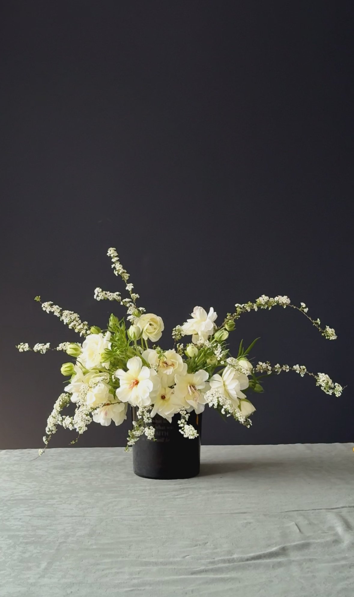 A bouquet of white flowers, including roses and other blooms, arranged in a black vase on a light-colored surface against a dark background. Stockholm high end event florist
