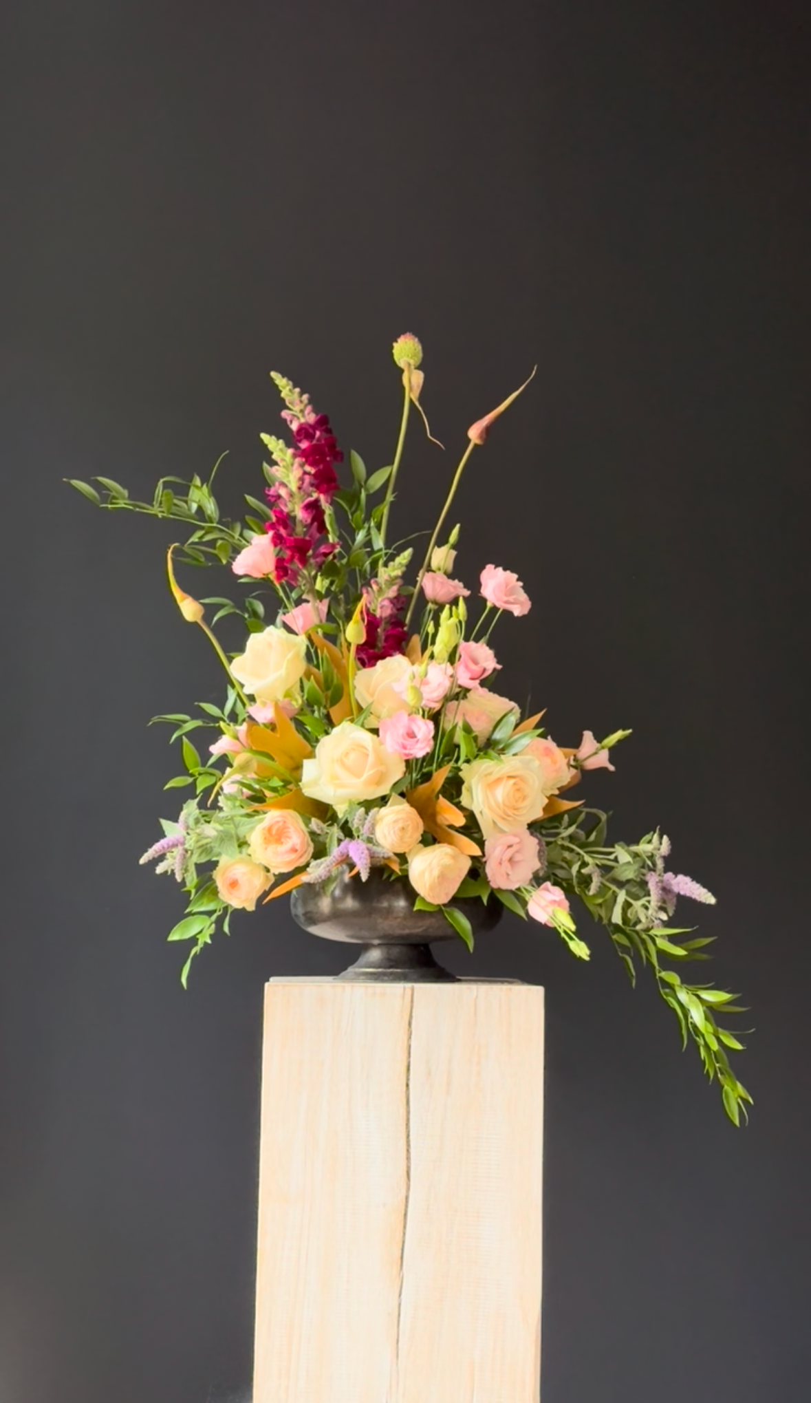 A floral arrangement in a dark metallic vase on a wooden pedestal, featuring pink roses, purple lavender, and various green foliage against a dark background. Stockholm high end event florist