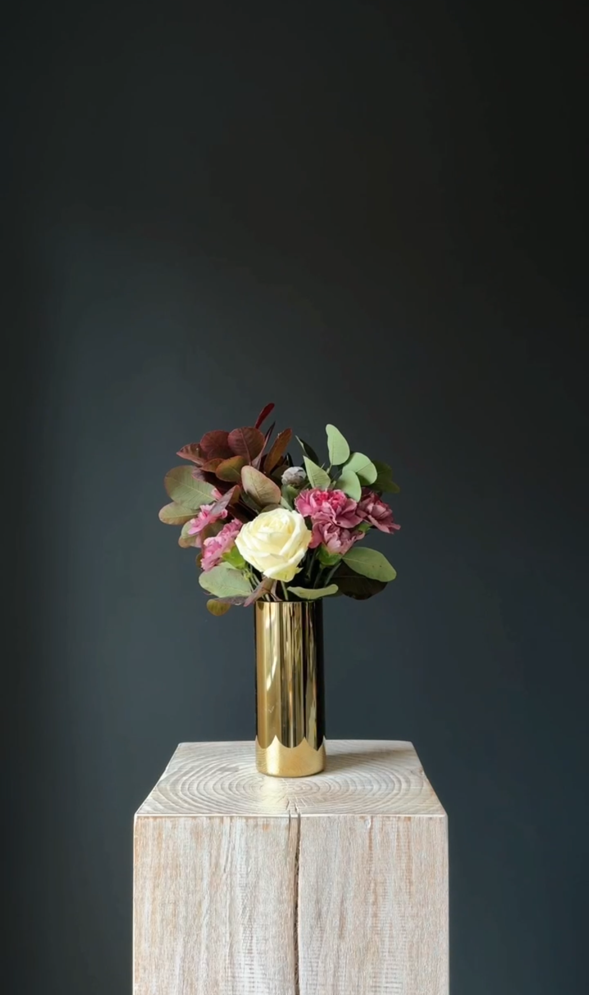 A bouquet of pink, cream, and green flowers in a tall, shiny gold vase, placed on a light wooden pedestal against a dark gray background. Stockholm high end event florist