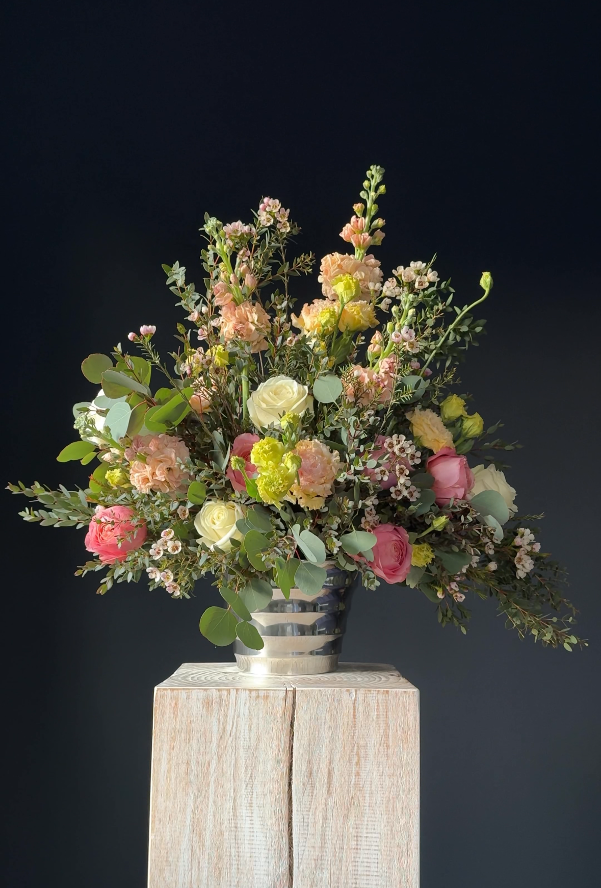 A colorful bouquet of flowers in a silver striped vase on a wooden pedestal against a dark background. Stockholm high end event florist