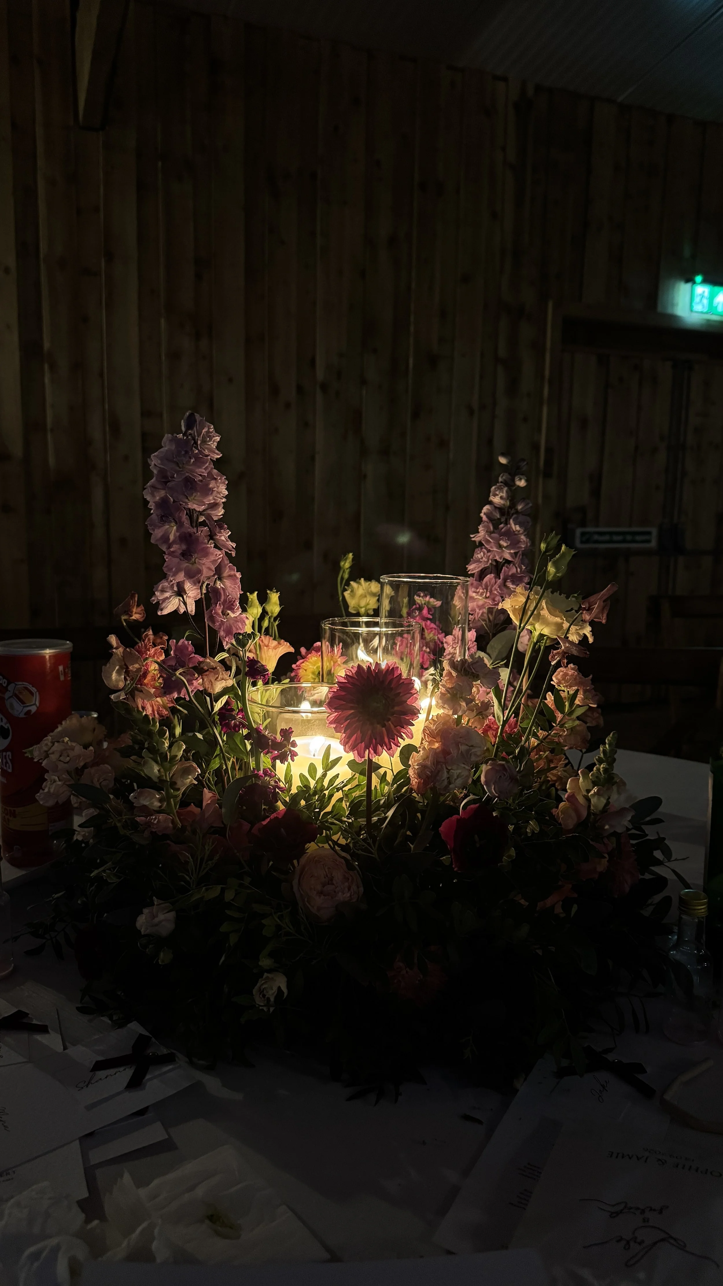 En blomsterarrangemang med ljus i glaslyktor, omgivna av rosa, lila och gula blommor. Bakgrund av trävägg och en nöddörrsskylt med grönt ljus.
