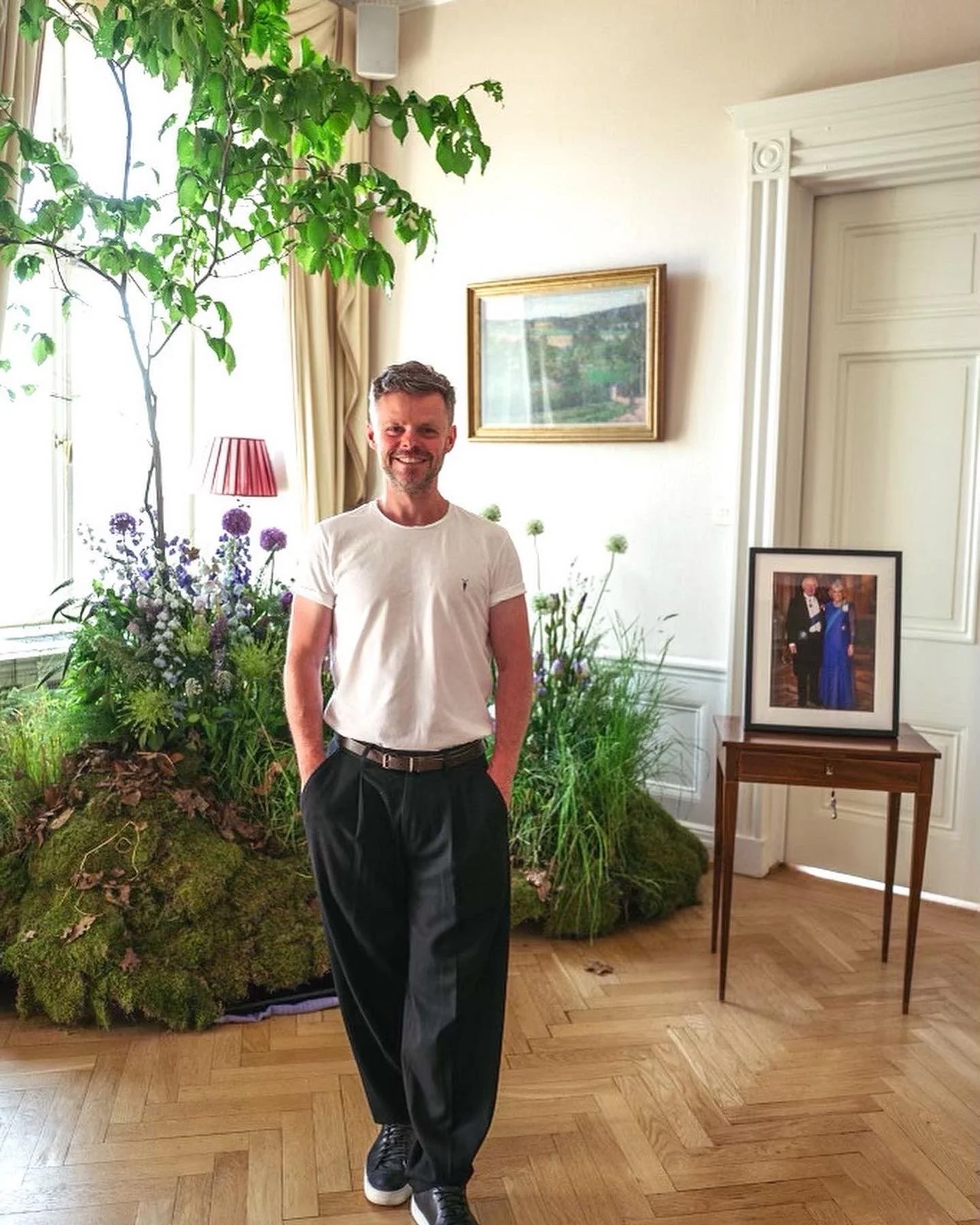 John stands indoors by a window and a garden display of flowers and moss. He wears a white t-shirt, black pleated trousers, and sneakers. Behind him, there is a framed photograph on a small wooden table, a picture on the wall, and lush green plants.