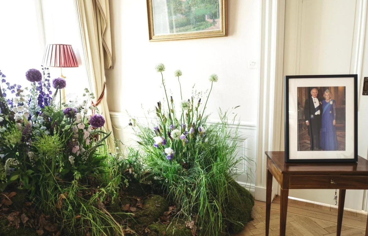 Indoor scene featuring a floral arrangement with purple and white flowers, a framed photograph of King Charles and the Queen