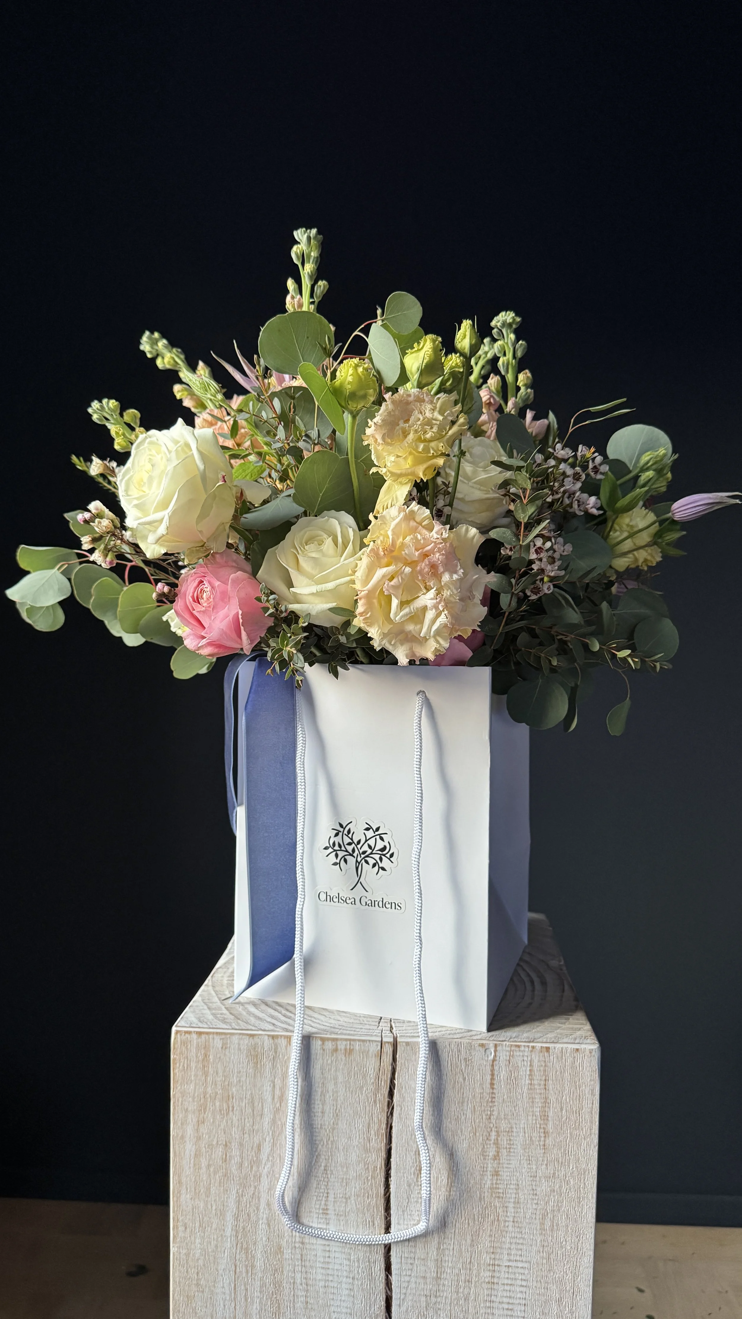 A bouquet of white, pink, and cream flowers with green leaves arranged in a Chelsea Gardens gift bag, placed on a wooden pedestal against a dark background. Stockholm high end event florist
