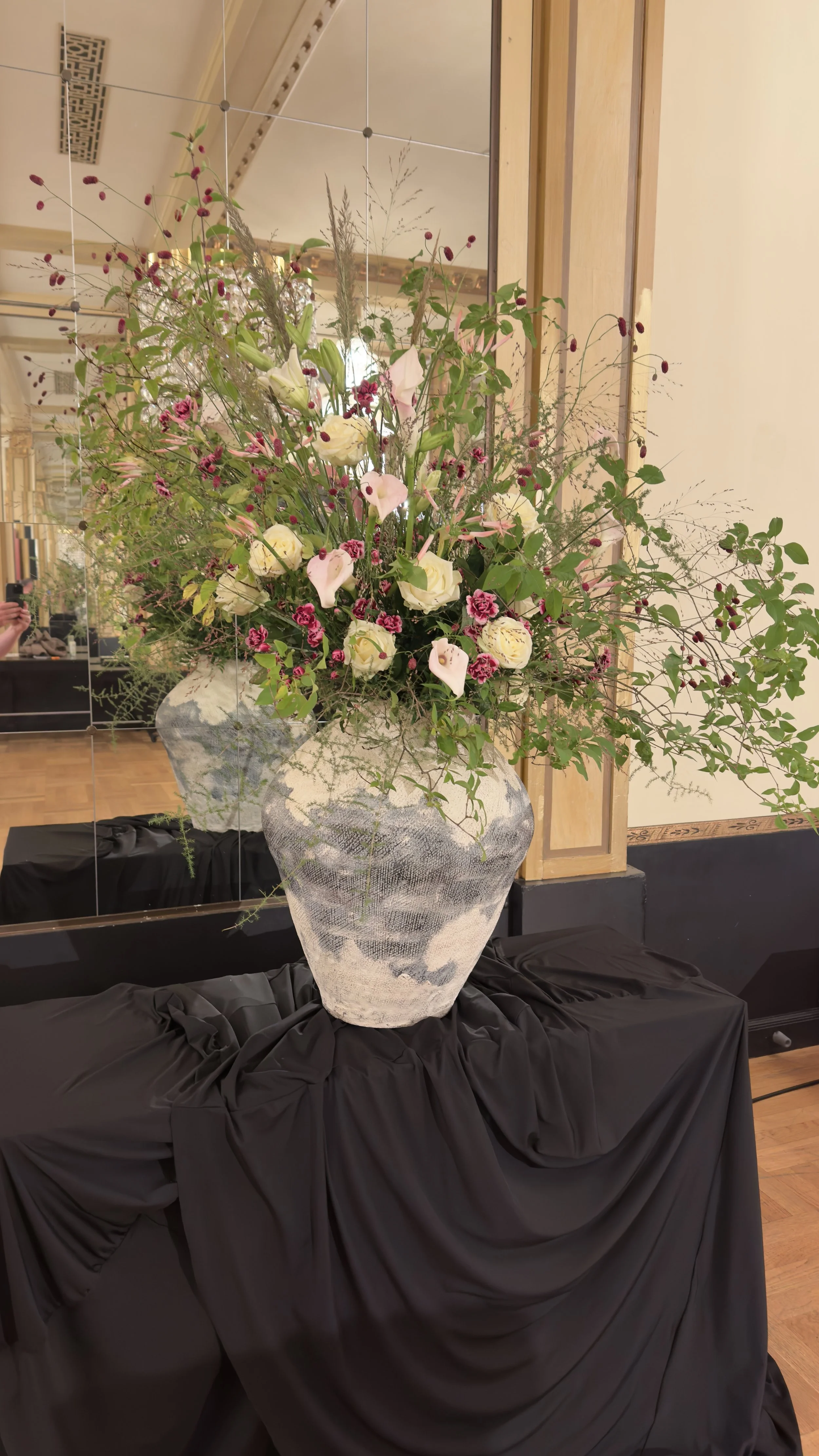 Large floral arrangement in a white and gray textured vase, placed on a black draped table in a room with wooden floors and mirrors. Stockholm high end event florist