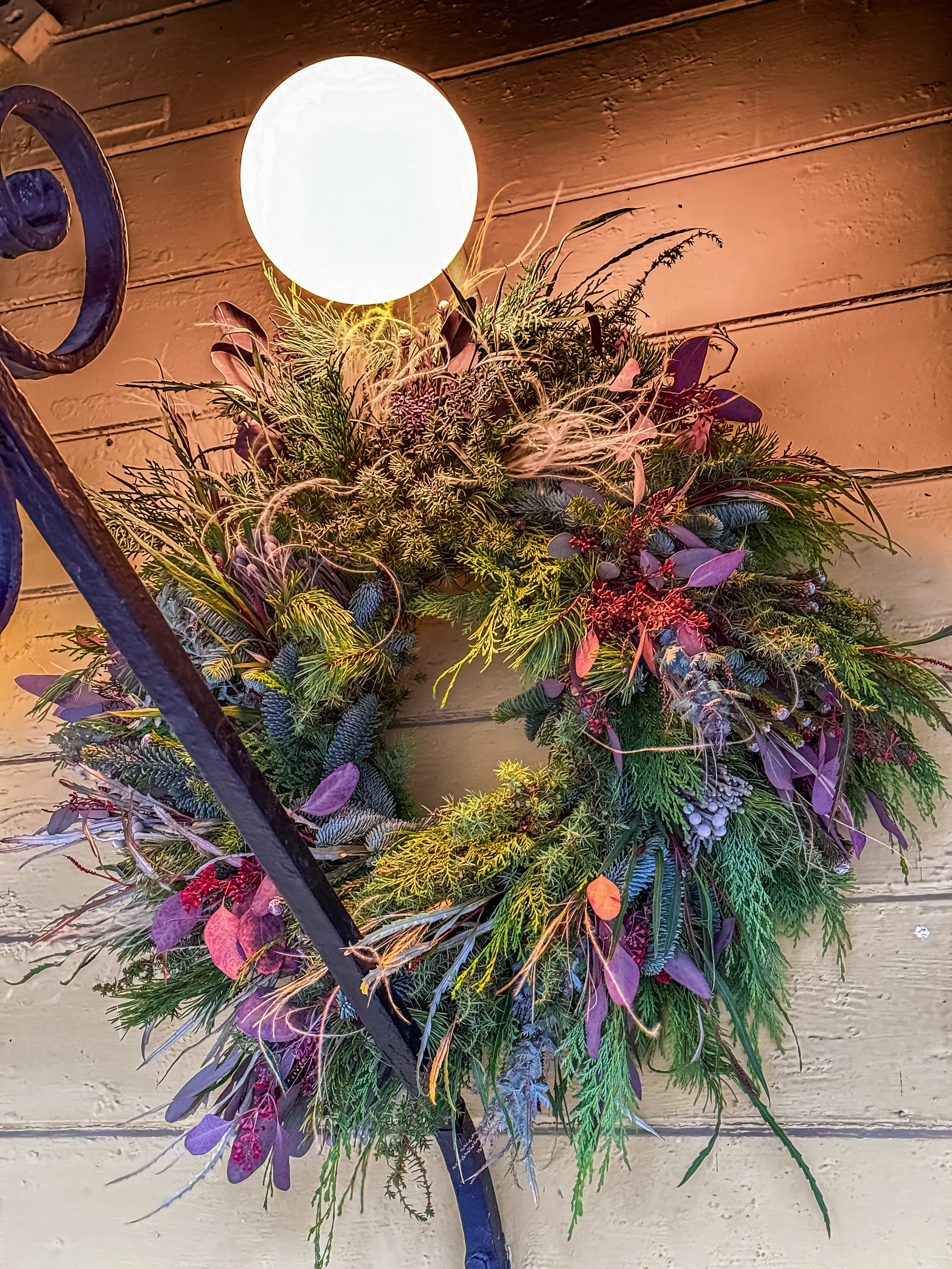 A decorative wreath made of various green and purple foliage, hanging below a round white light fixture on a wooden wall. Stockholm high end event florist