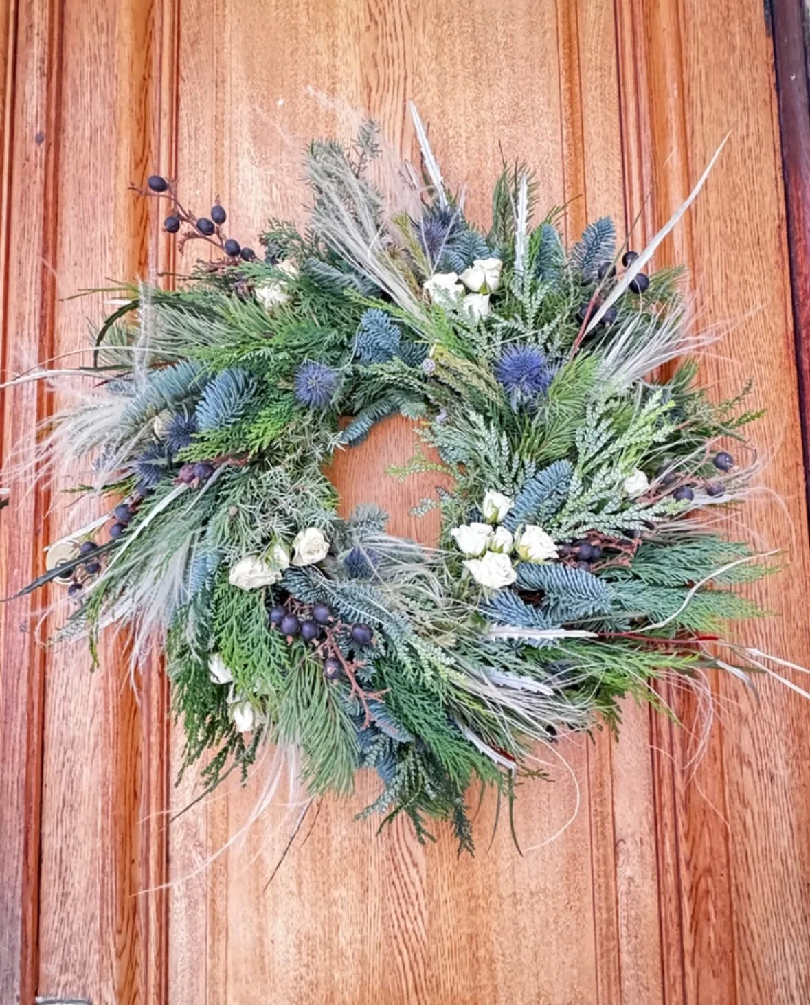 A Christmas wreath made of various green, blue, and white foliage, including conifer branches, eucalyptus, and decorative berries, hung on a wooden door. Stockholm high end event florist