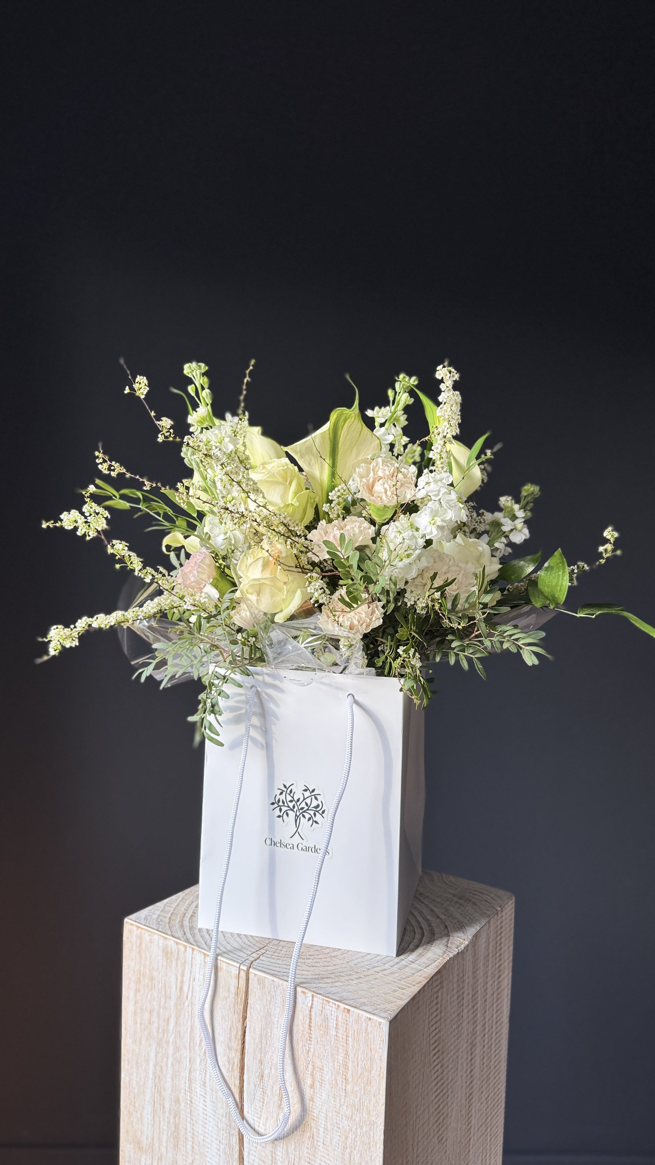 A white shopping bag with flowers inside, placed on a wooden pedestal against a black background. Stockholm high end event florist