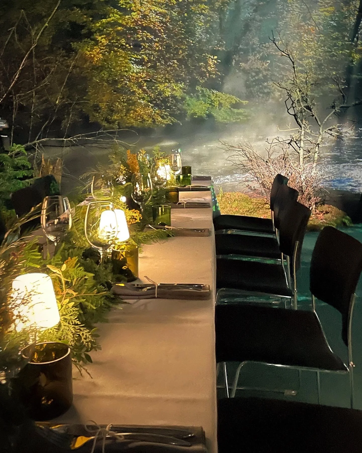 A beautifully set banquet table with candles and greenery, overlooking a misty river at night surrounded by trees.