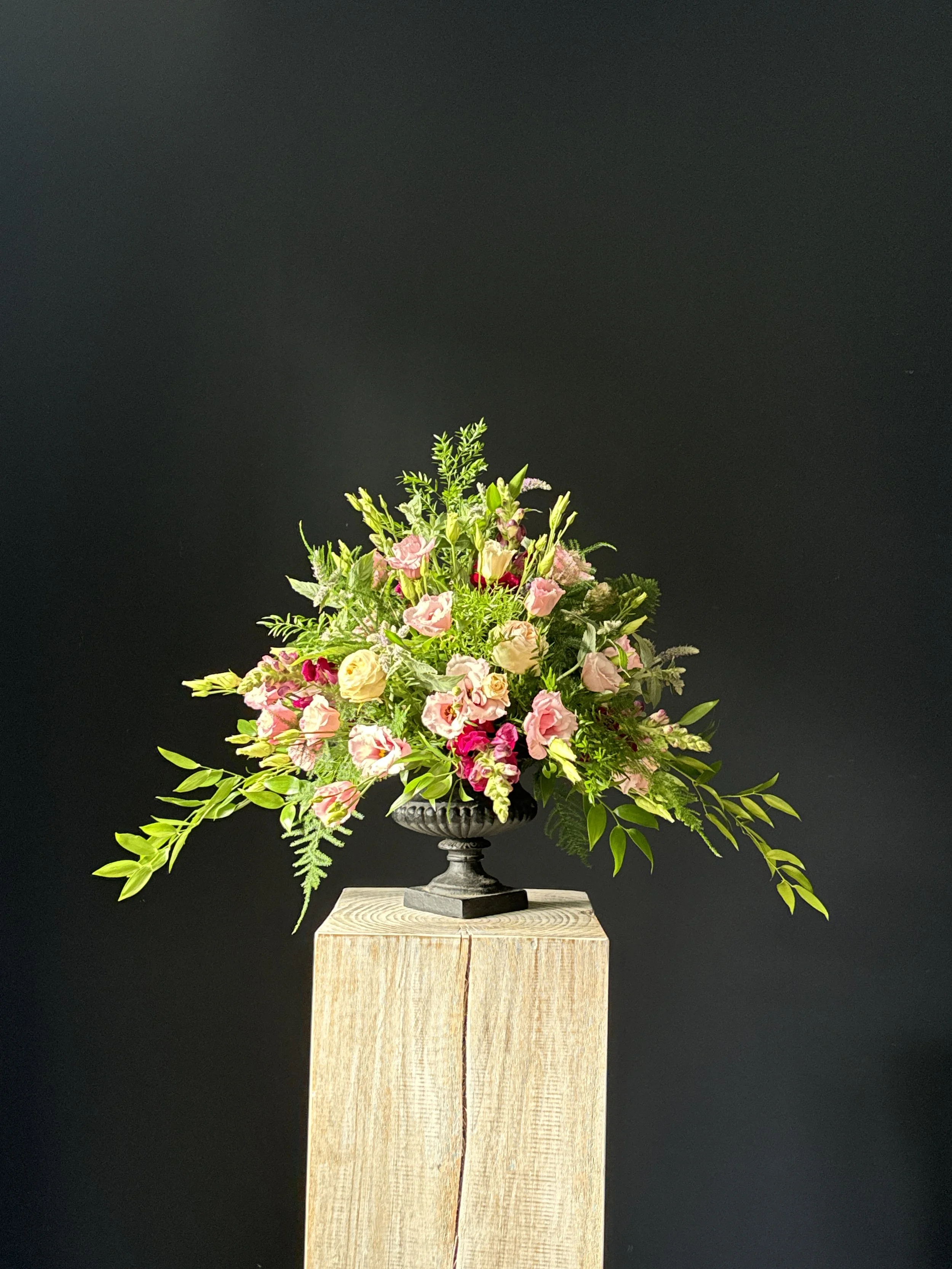 A colorful floral arrangement with pink, white, and red flowers in a black vase on a light wooden pedestal against a black background. Stockholm high end event florist