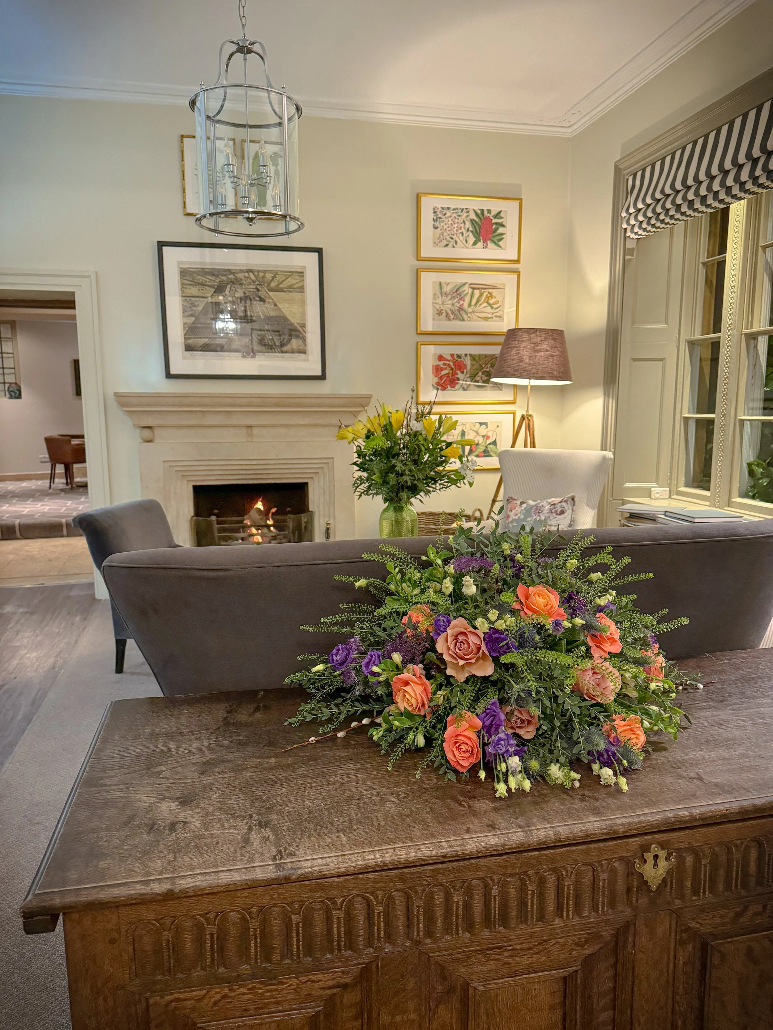 A living room with a wooden table in the foreground holding a large colorful flower arrangement. In the background, a fireplace with a roaring fire, a white armchair, a tall floor lamp, and framed botanical prints on the wall. Large windows with stri