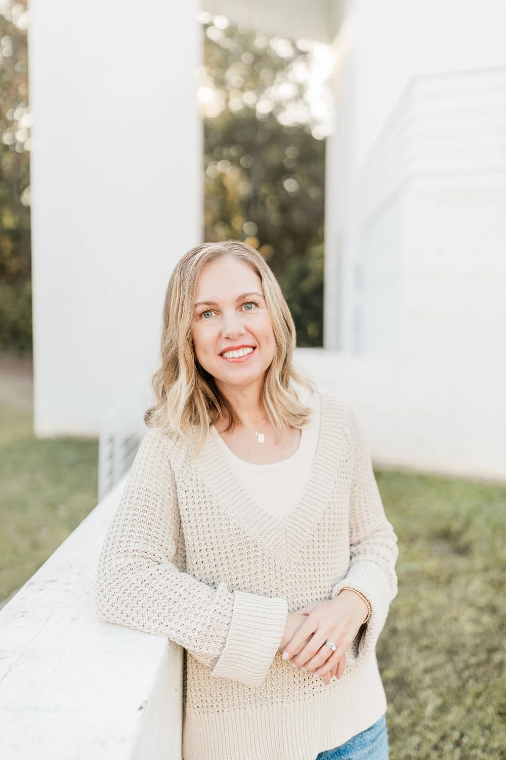 A woman with shoulder-length blonde hair, blue eyes, and a light complexion smiling and leaning against a white wooden railing outdoors. She is wearing a beige knitted sweater over a white top, with a gold bracelet and necklace, and has a wedding ring on her left hand.
