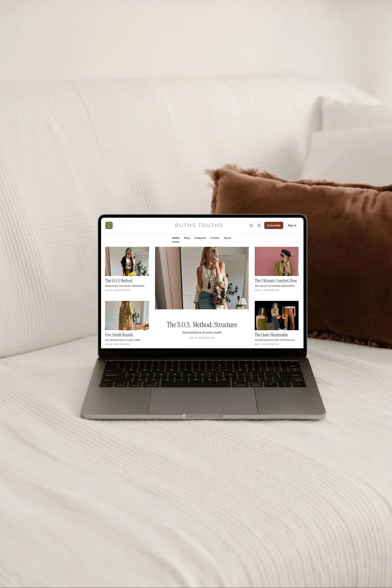 A laptop on a white textured table shows a fashion blog website named Ruths Truths with articles and images of women in stylish clothes, against a background of a white sofa with brown pillows.
