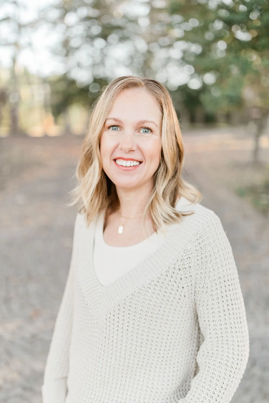 A woman with blonde hair and blue eyes smiling outdoors, wearing a light-colored sweater and a necklace, with blurred trees and natural scenery in the background.