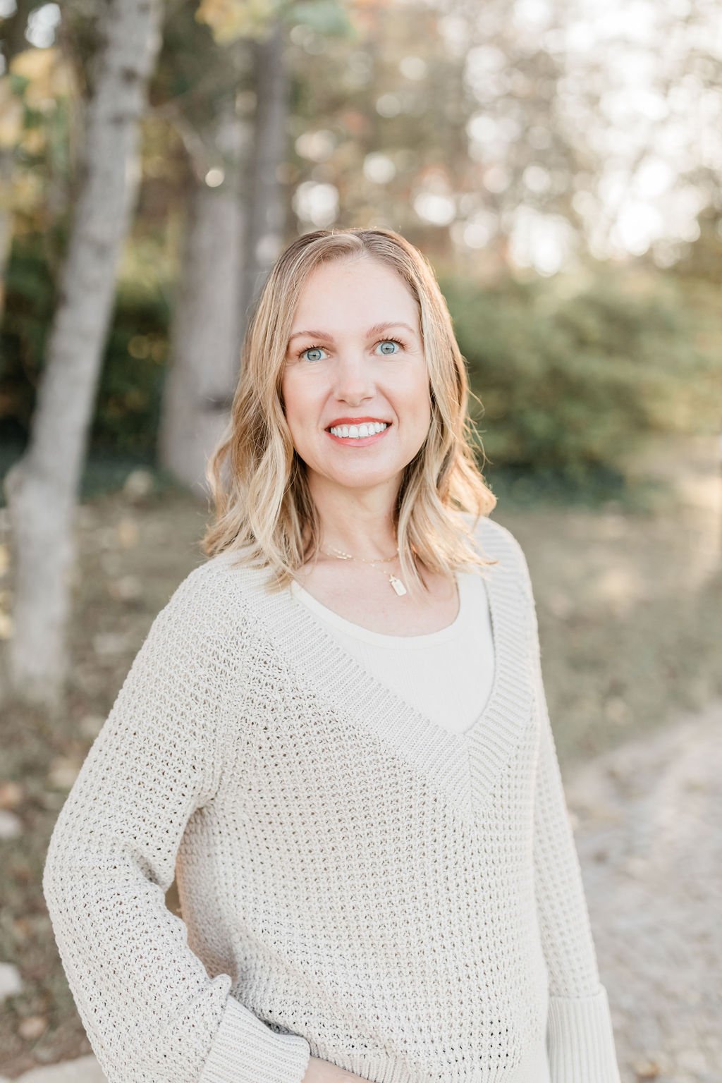 A woman with blonde, wavy hair and blue eyes smiling outdoors in front of trees with autumn leaves, wearing a cream-colored sweater and a white top.