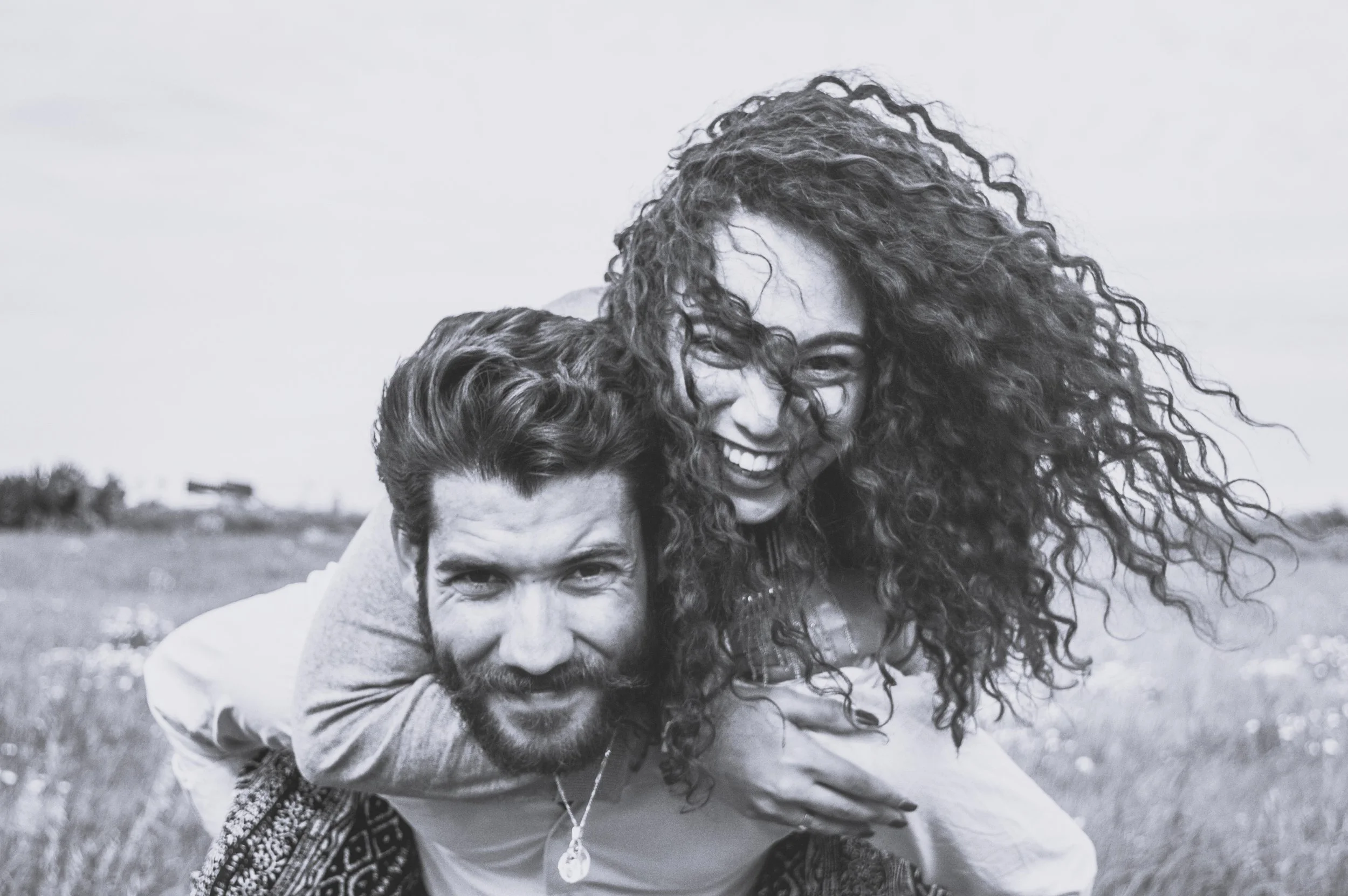 A black and white photo of a man and woman outdoors in a field, smiling and having fun. The woman is on the man's back, playfully tousling her curly hair.