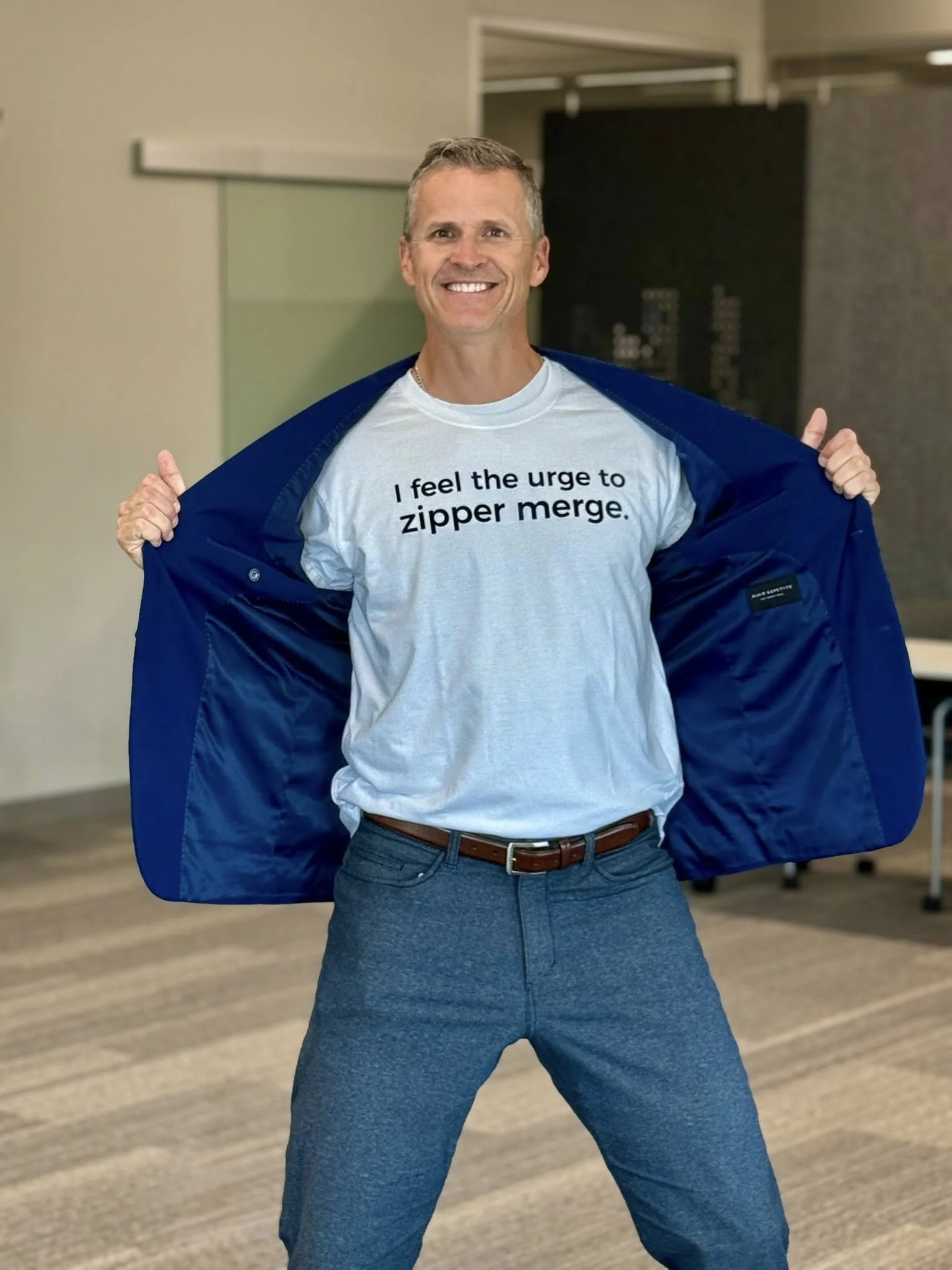 A man smiling and posing with an open blue jacket revealing a white t-shirt that says 'I feel the urge to zipper merge.' in black text.
