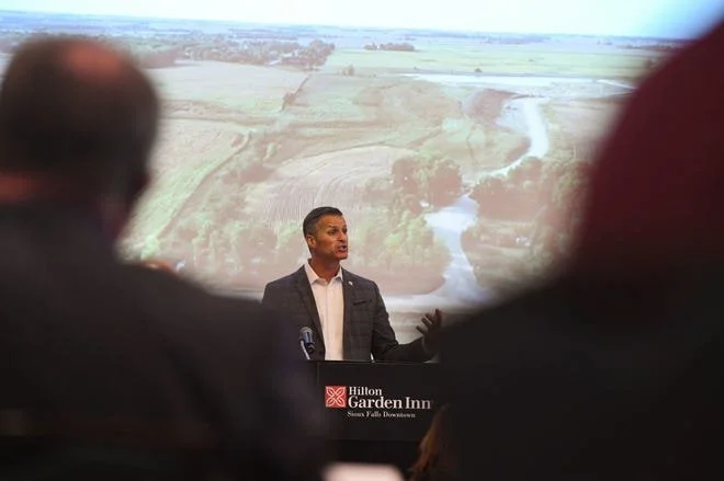A man in a suit is speaking at a conference with a projected landscape of fields, a river, and farmland behind him. The conference is held at Hilton Garden Inn.