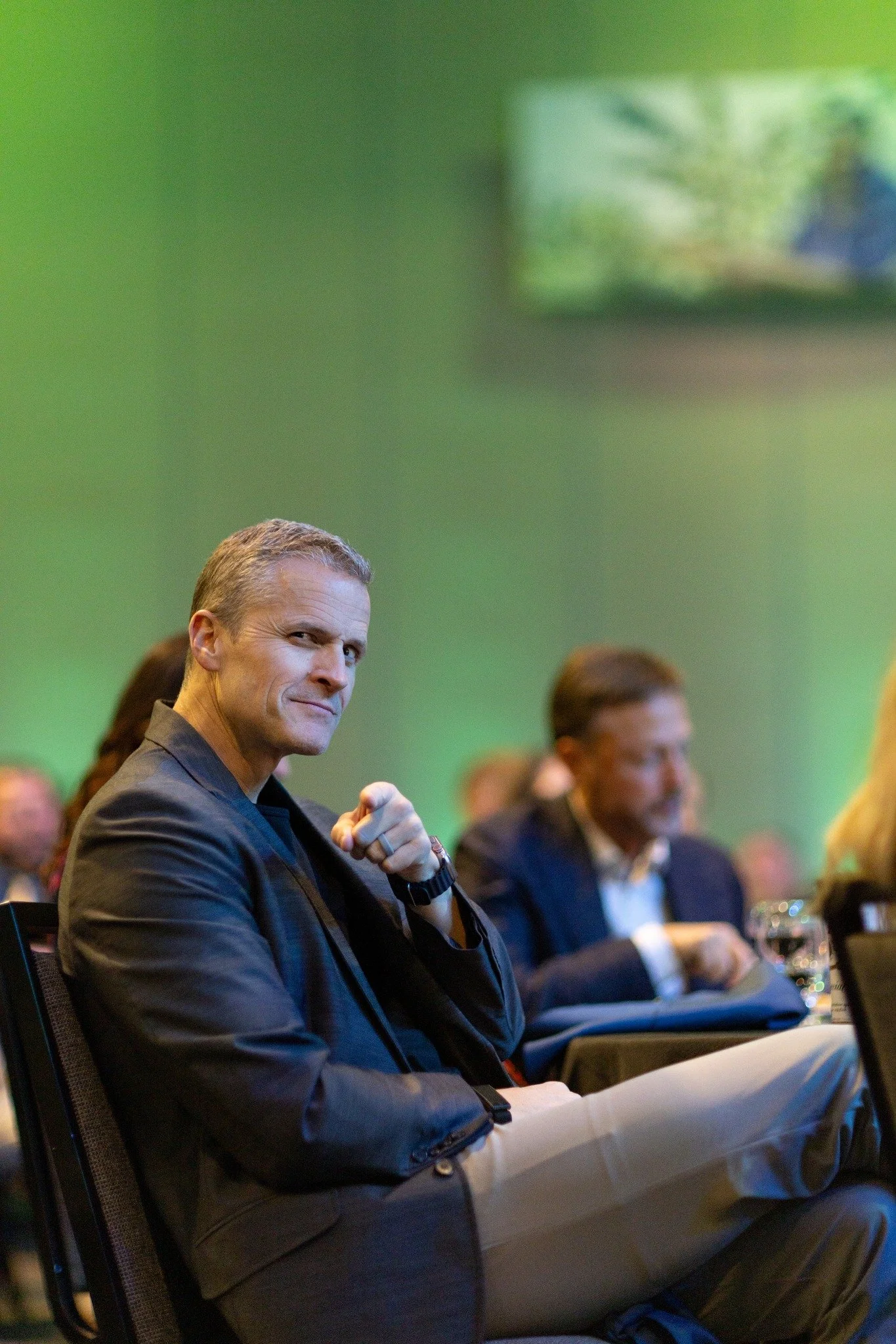A man in a suit sitting at a conference, looking at the camera with a confident expression, with other attendees in the background.