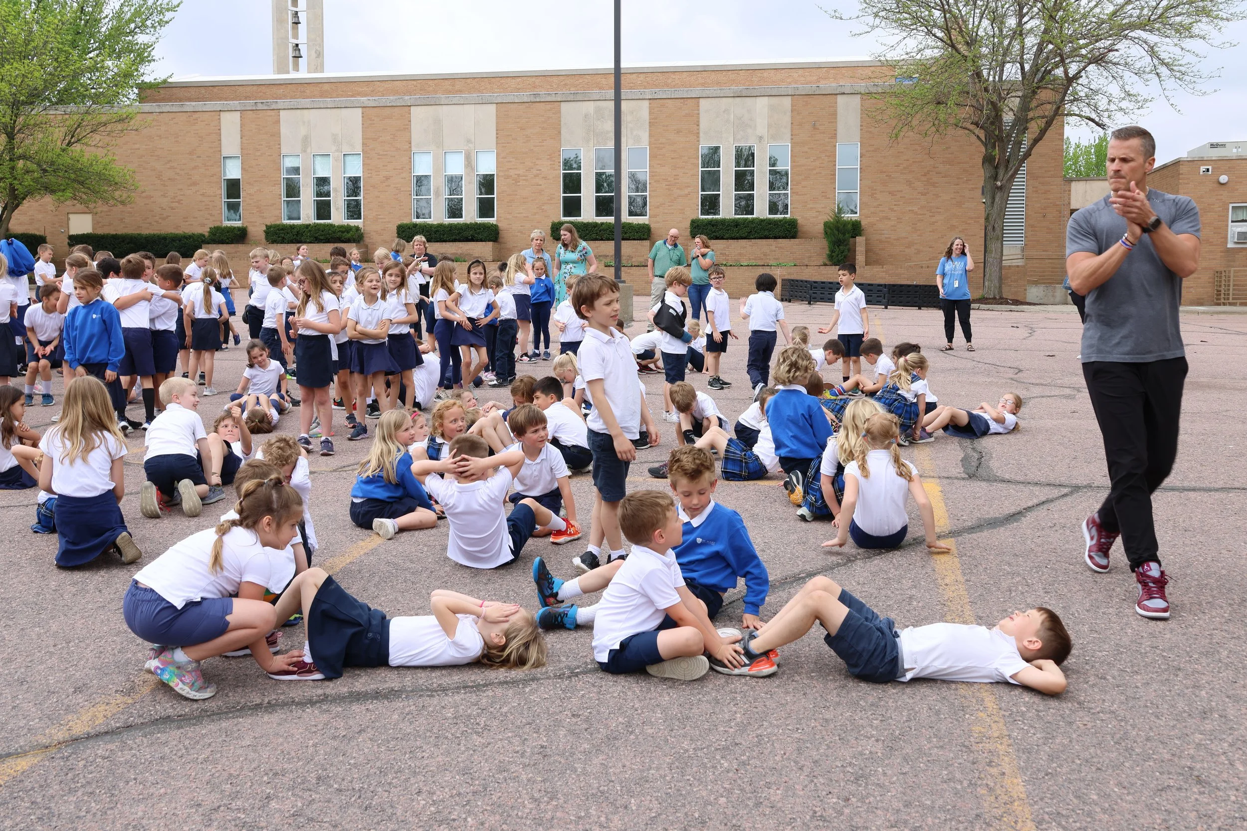 A group of elementary school children in uniform are participating in an outdoor activity on a school playground, with some sitting or lying on the ground and others standing while a man walks past.