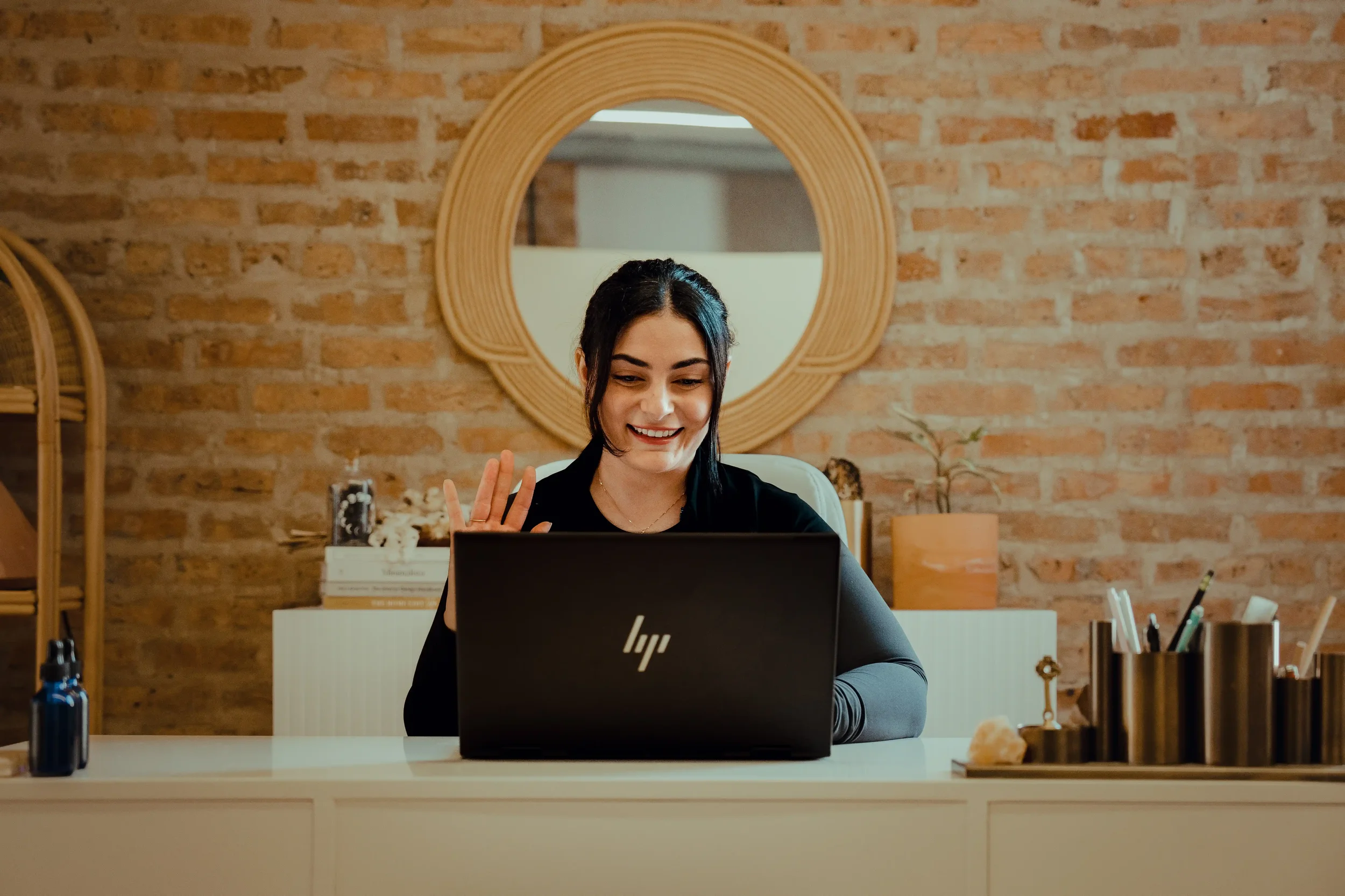 A PT doing a virtual session with a patient, the dark-haired woman is sitting in front of her laptop, waving hello.