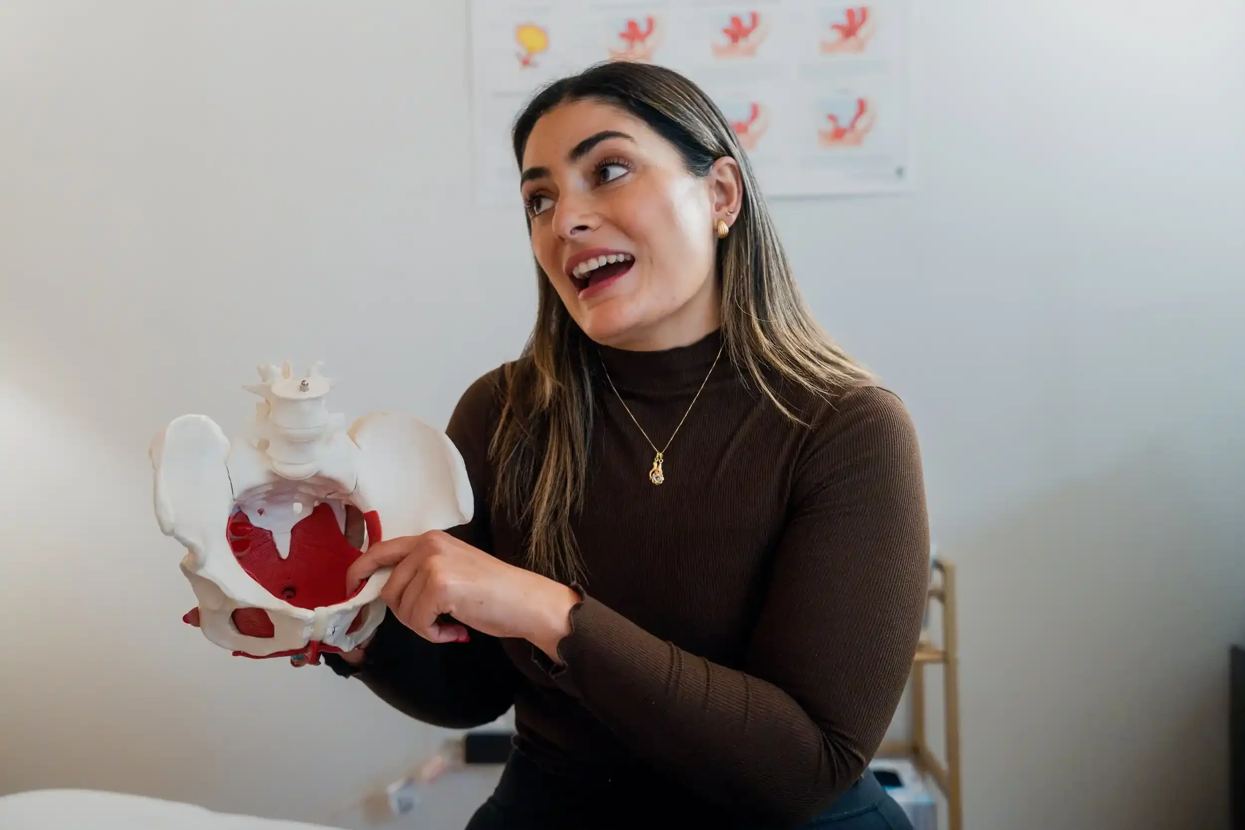 A woman holding a model of the human pelvis, the model shows bones and muscle structure, in a cozy office environment.