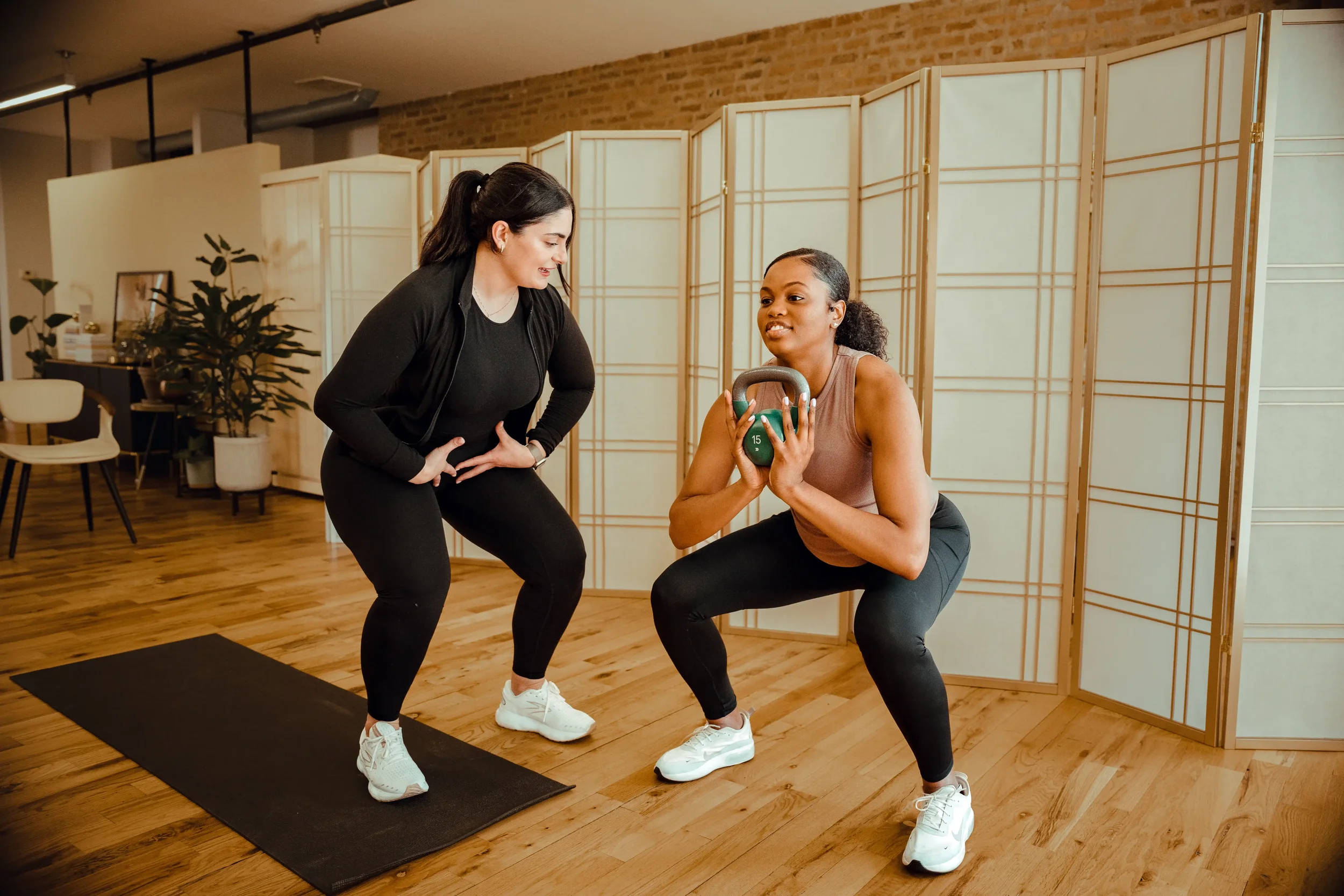 Woman holding a kettlebell squatting in a bright and inviting space while a physical therapist cues her.